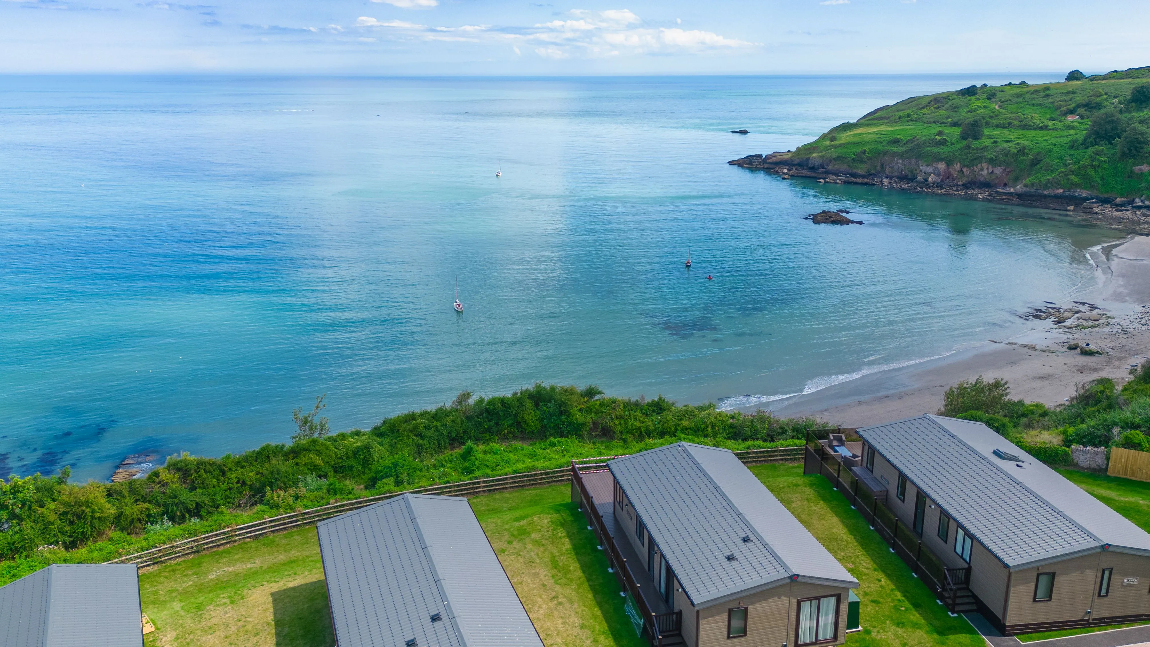 Coastal bay with calm blue sea, sandy beach, green headland and holiday lodges in the foreground