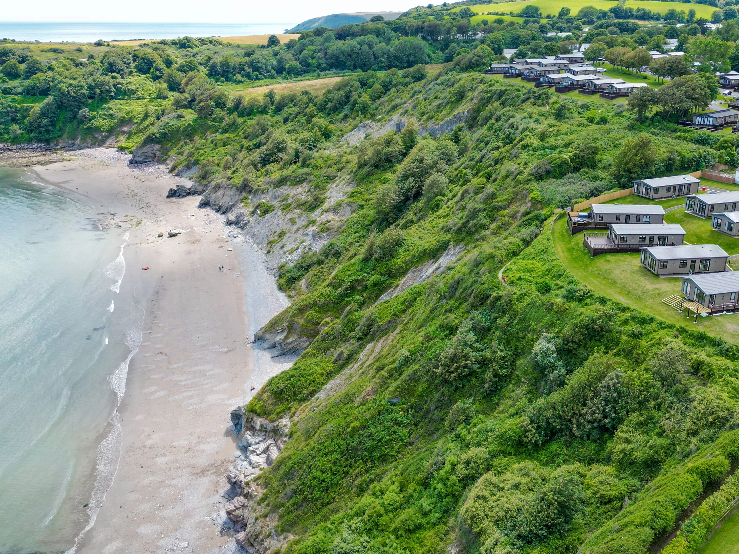 Aerial view of a coastal holiday park on green cliffs beside a sandy beach and calm sea