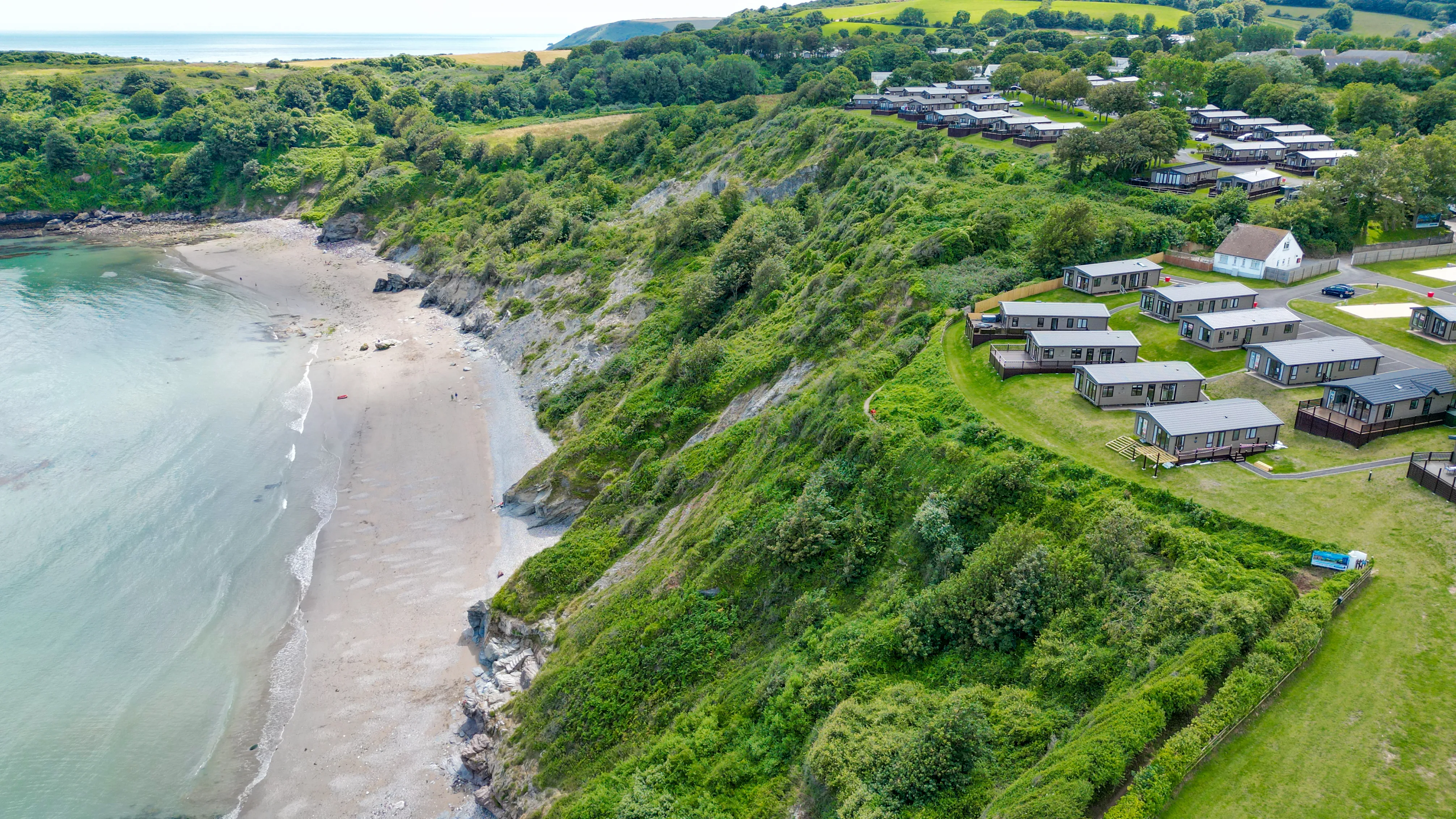 Aerial view of a coastal holiday park on green cliffs beside a sandy beach and calm sea