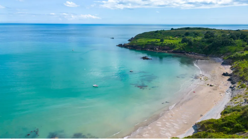 Aerial view of a turquoise bay with sandy beach, anchored boats and green headland under a blue sky
