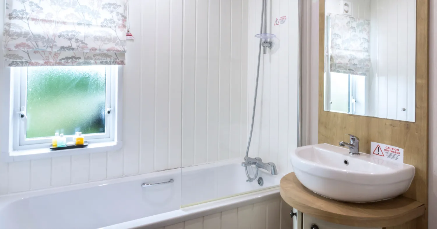 Bright white bathroom with a bath and overhead shower, window with patterned blind, and a washbasin with mirror