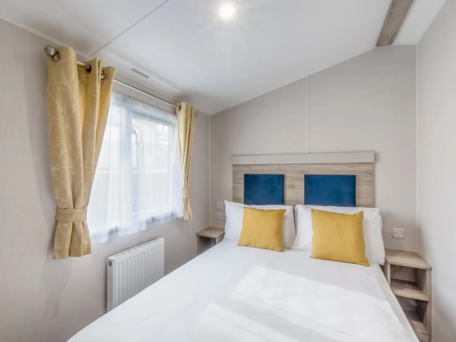 Bright double bedroom with white bedding, blue headboard panels and yellow cushions beside a window with yellow curtains.