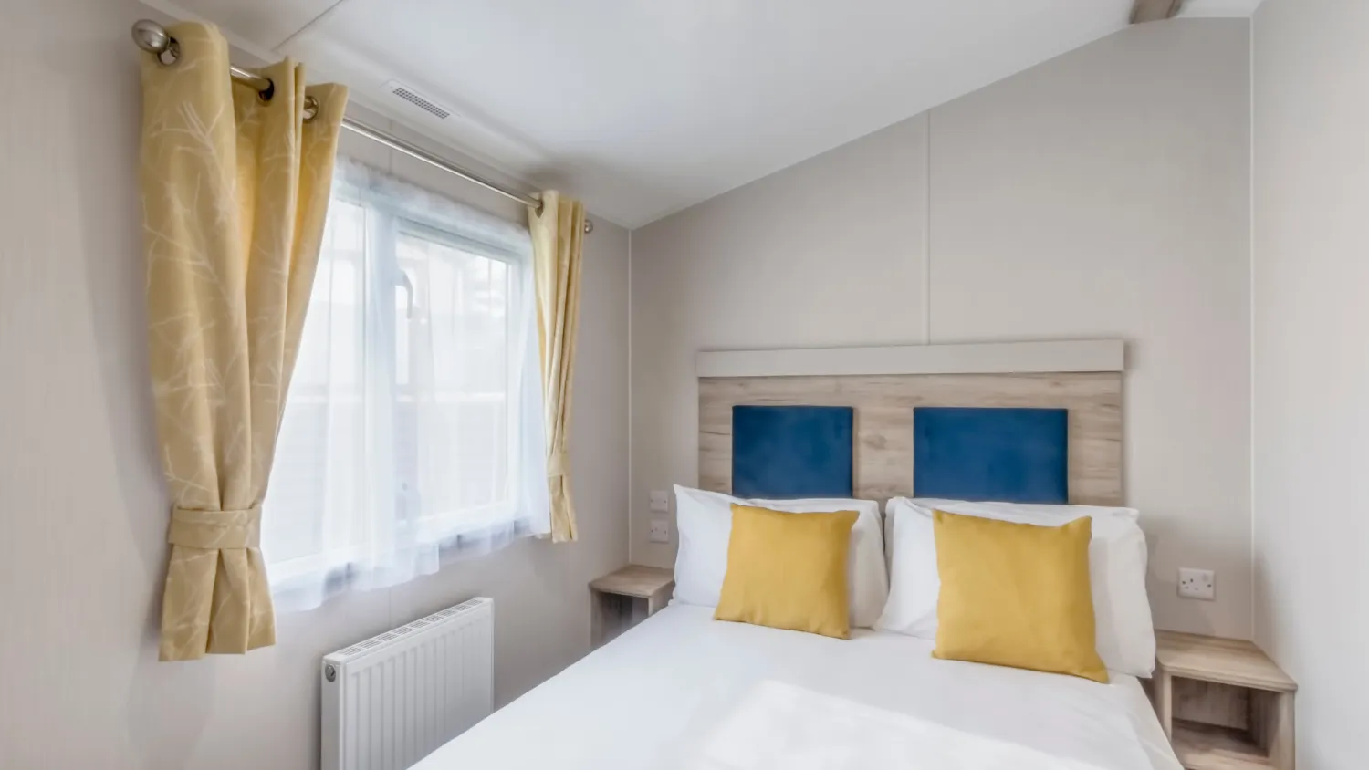 Bright double bedroom with white bedding, blue headboard panels and yellow cushions beside a window with yellow curtains.
