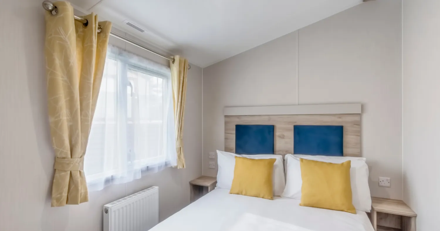 Bright double bedroom with white bedding, blue headboard panels and yellow cushions beside a window with yellow curtains.