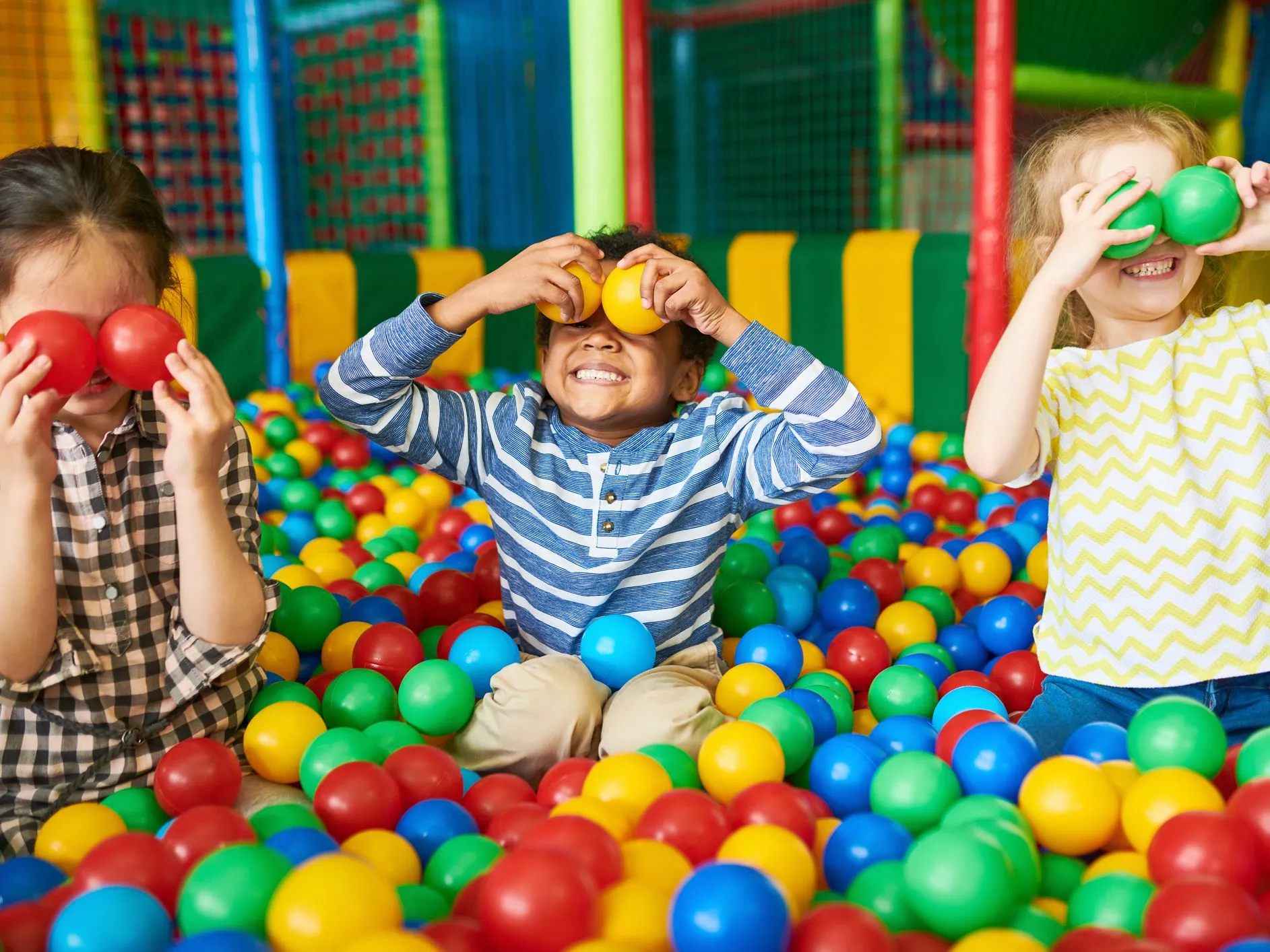 Three children playing in a colourful ball pit, holding plastic balls over their eyes