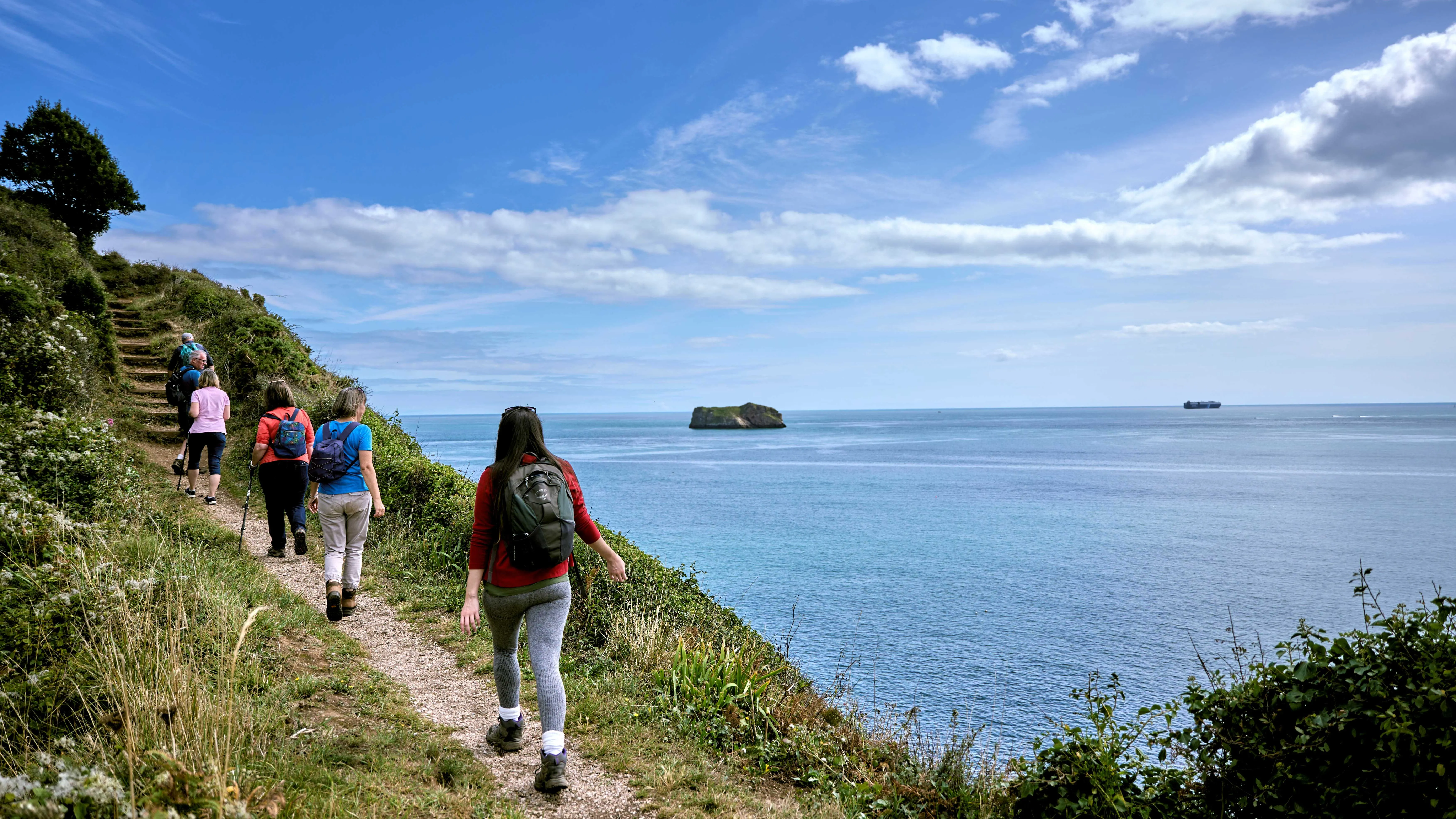 Walkers with backpacks on a coastal cliff path overlooking the sea under a blue sky