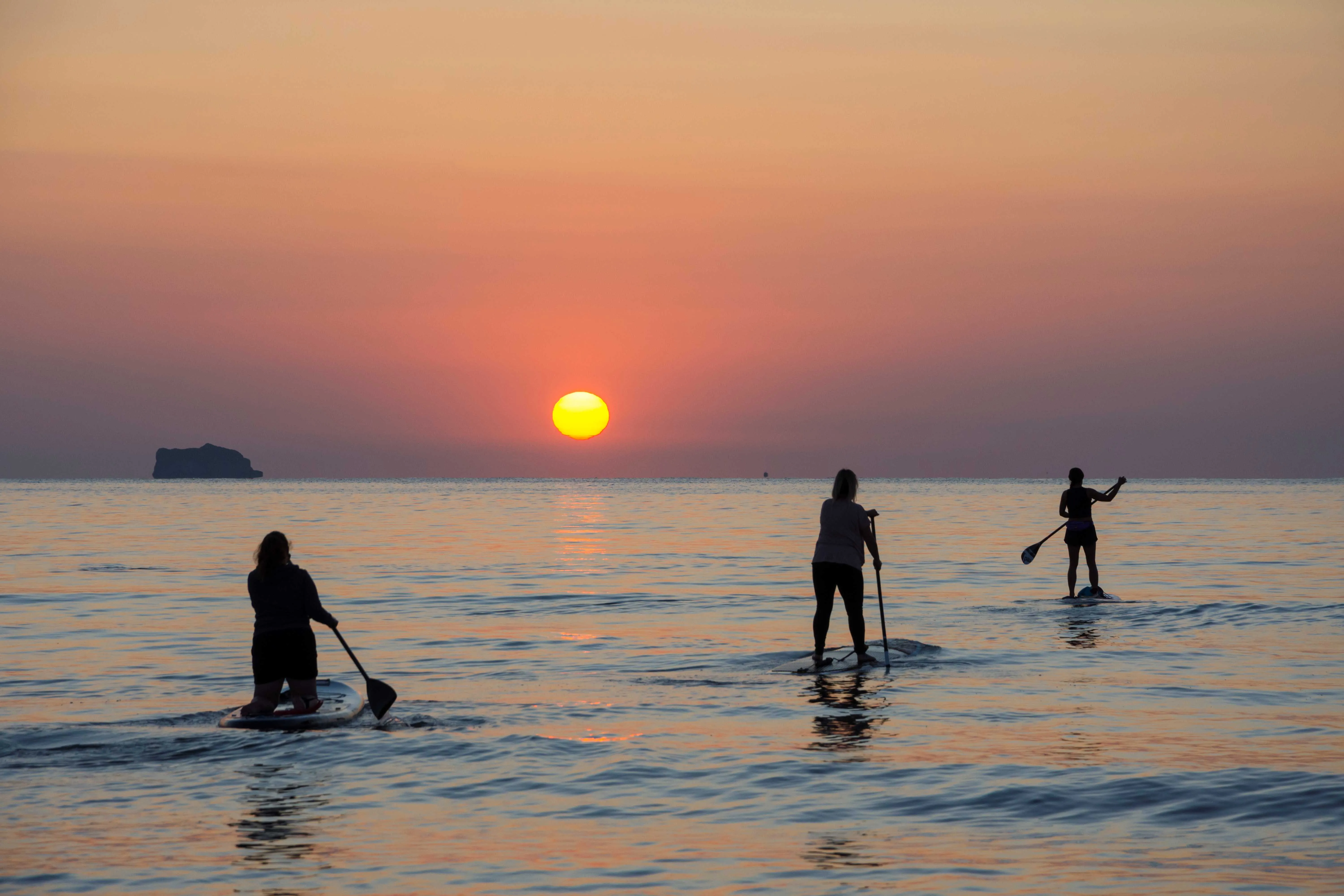 Silhouetted paddleboarders on calm sea at sunset with an island on the horizon