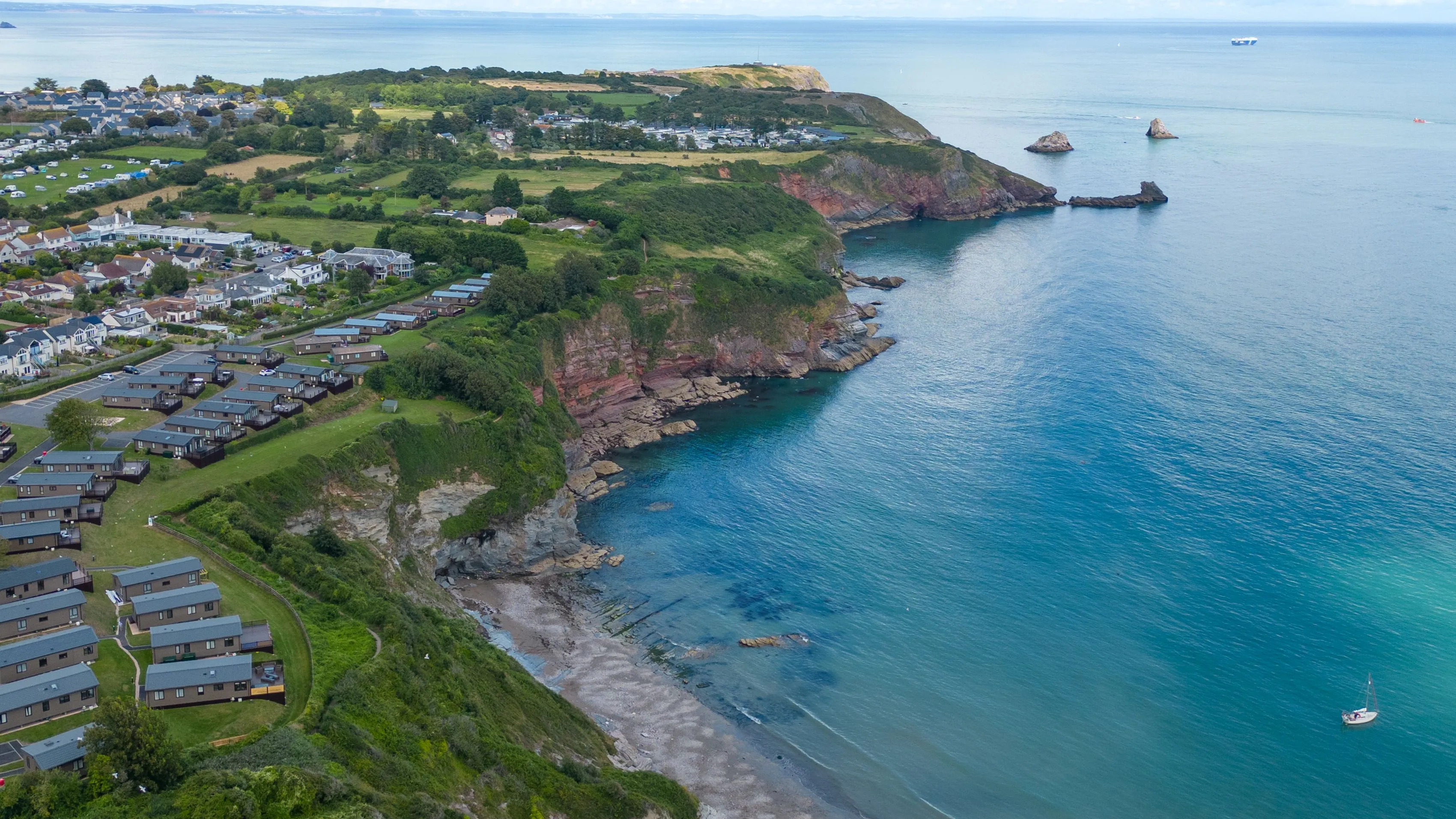 Drone view of Riviera Bay holiday park on a clifftop overlooking a sheltered bay with turquoise sea and rocky coastline.