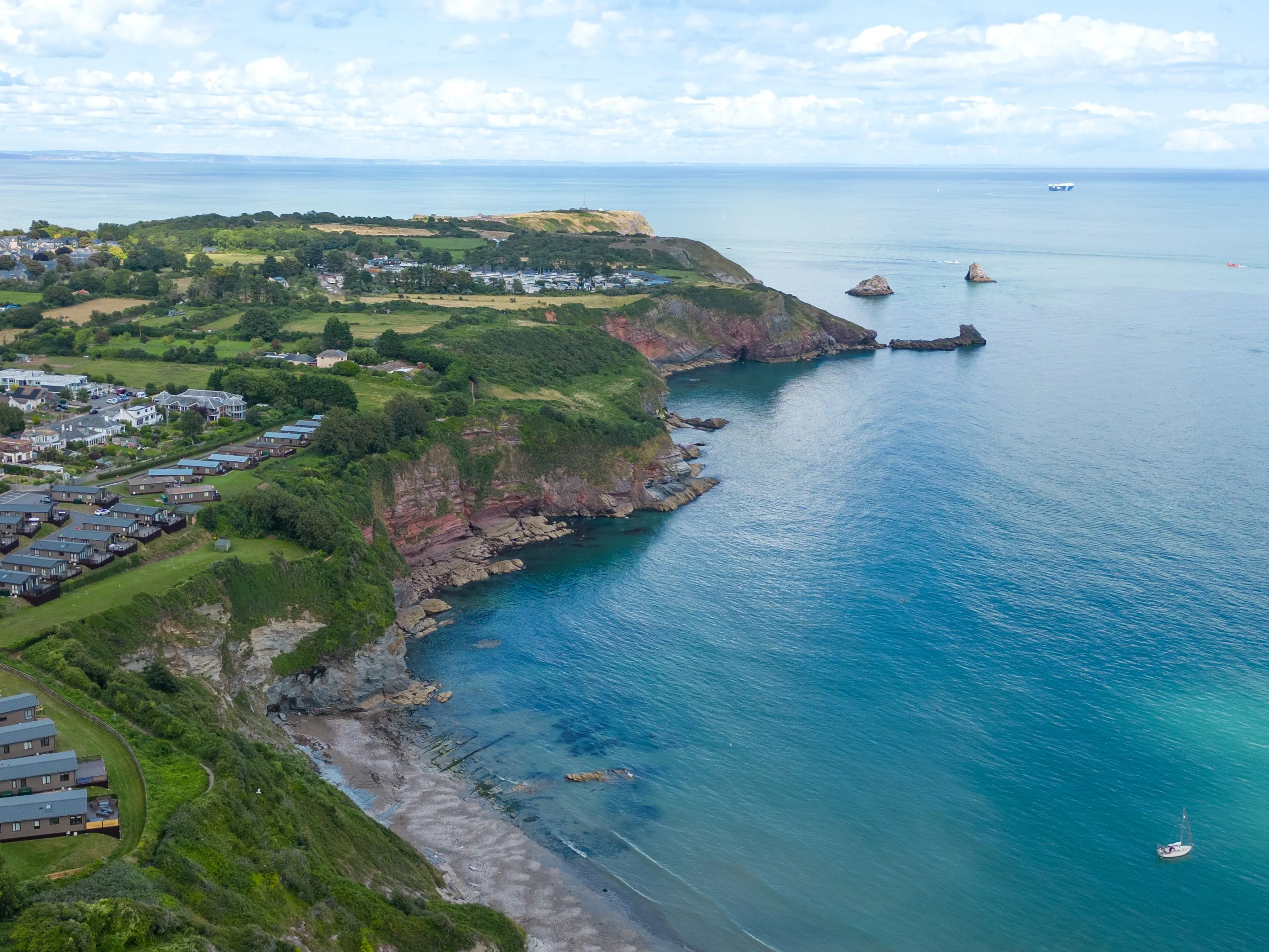 Drone view of Riviera Bay holiday park on a clifftop overlooking a sheltered bay with turquoise sea and rocky coastline.