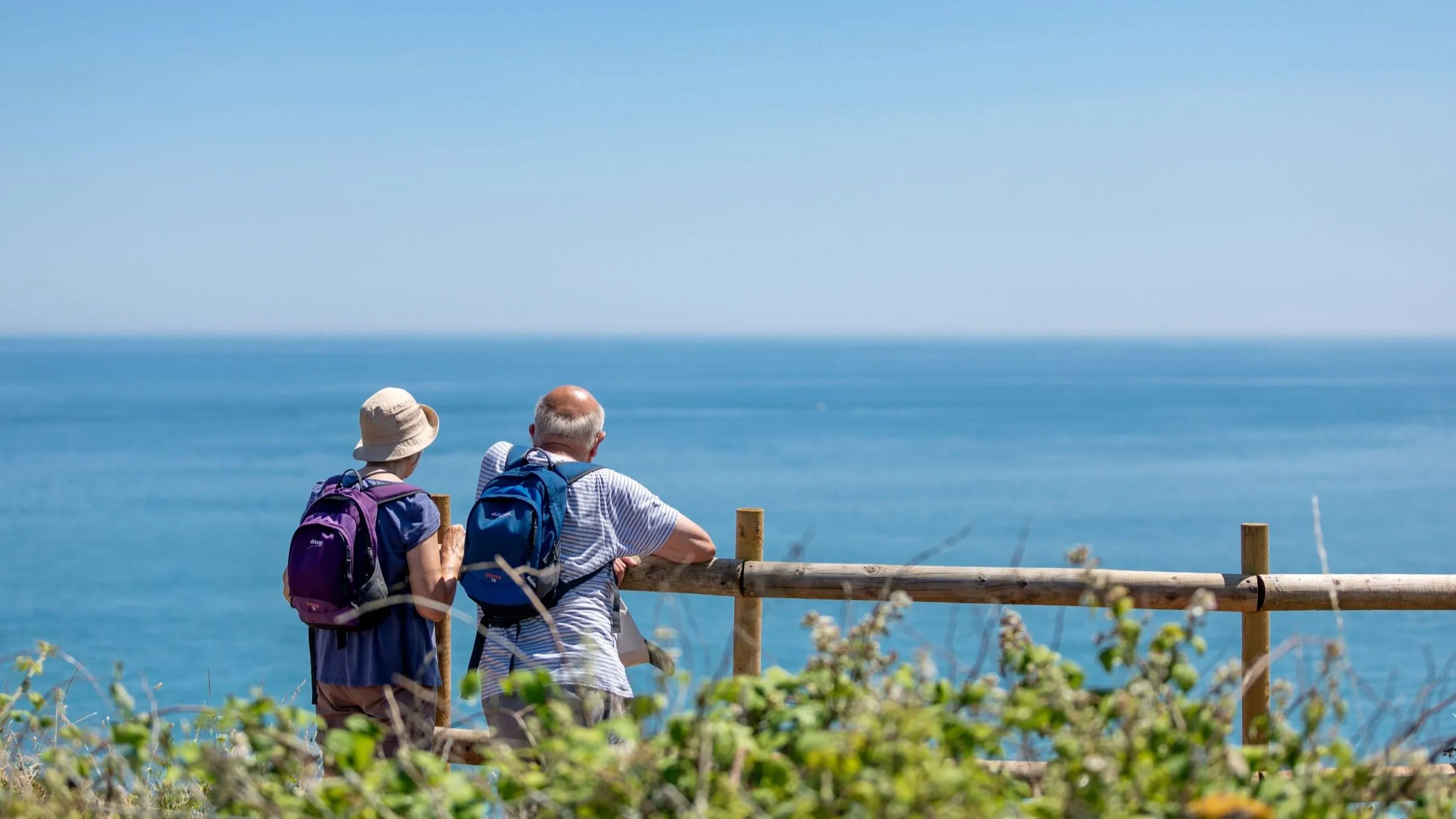 Two walkers with backpacks stand by a wooden fence overlooking the sea on a sunny day.