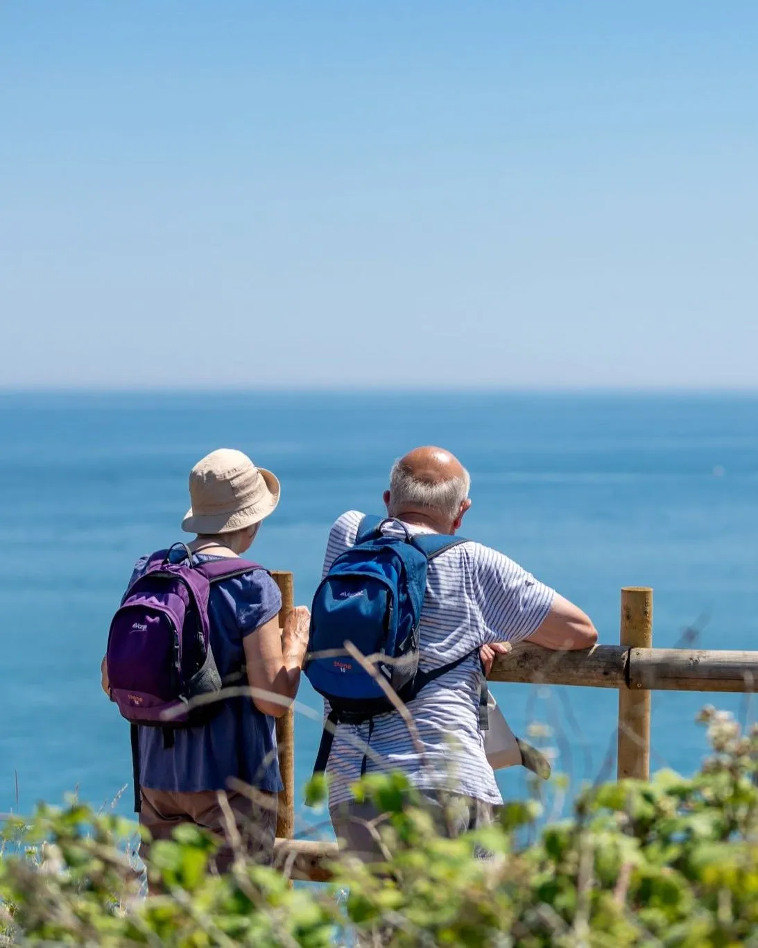 Two walkers with backpacks stand by a wooden fence overlooking the sea on a sunny day.