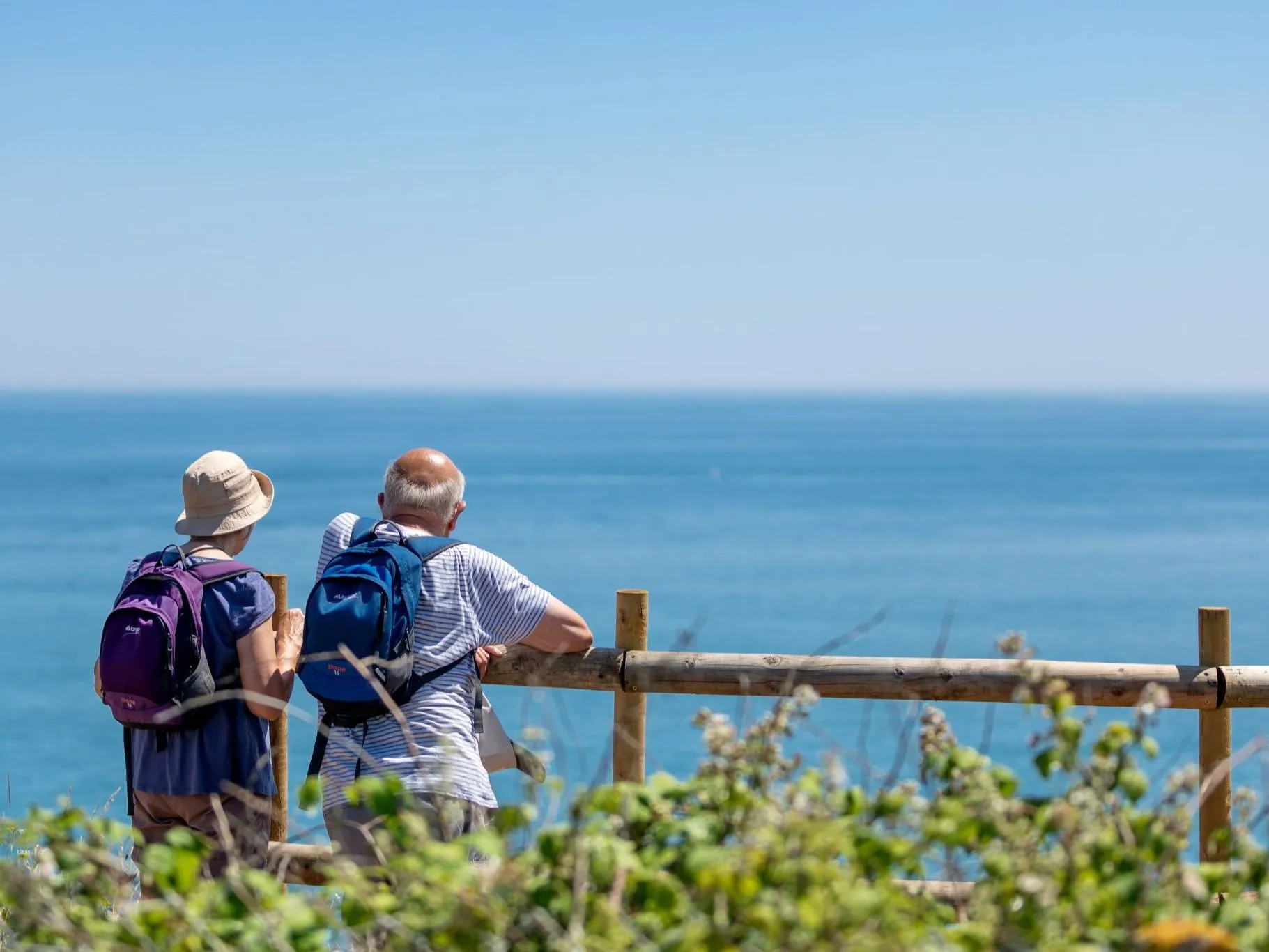 Two walkers with backpacks stand by a wooden fence overlooking the sea on a sunny day.