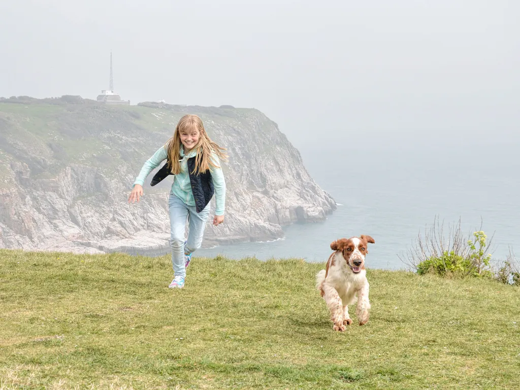 Young girl running with a dog on the clifftop at Berry Head on the South West Coast Path, with the sea and rocky cliffs behind.