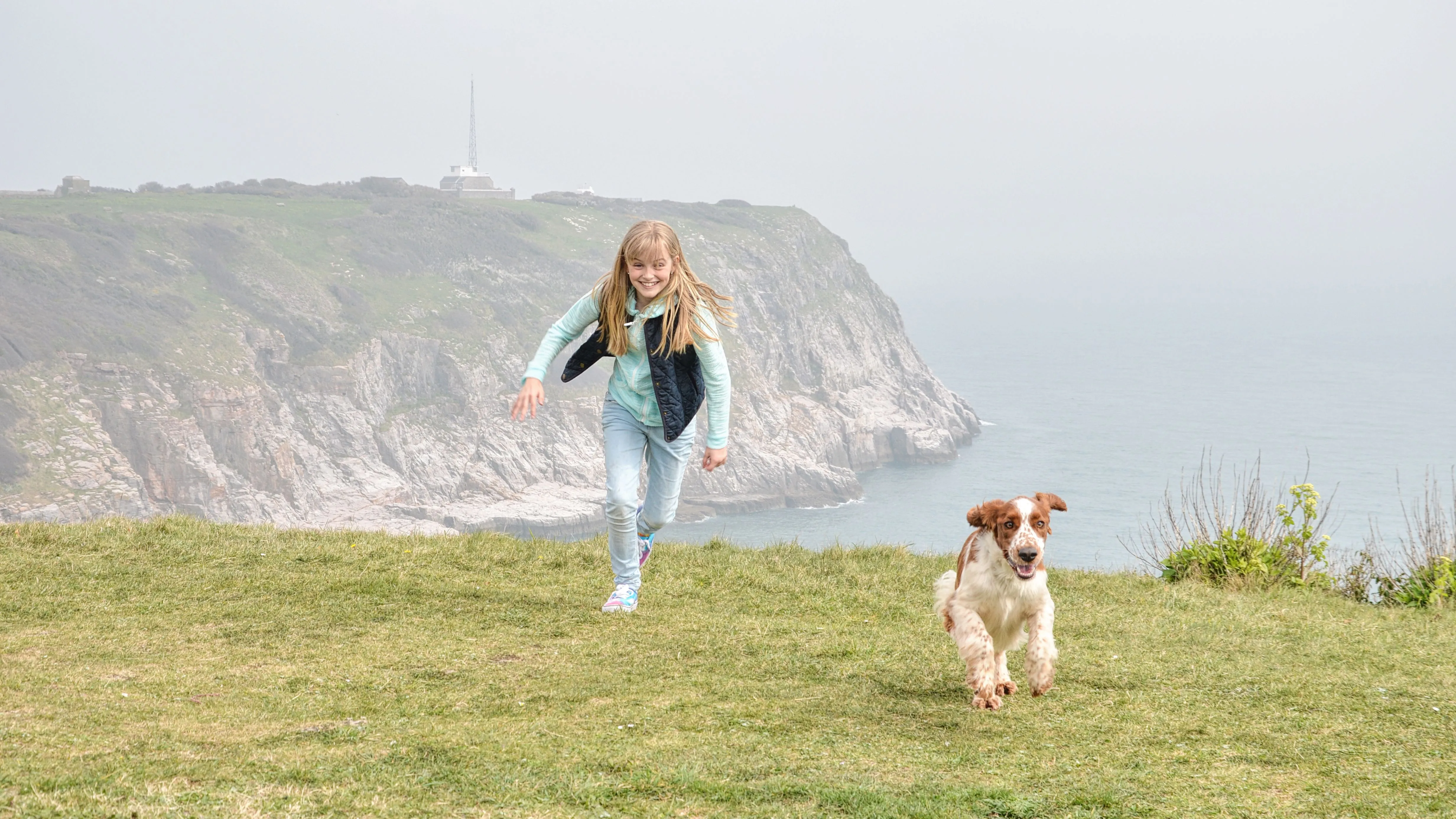Young girl running with a dog on the clifftop at Berry Head on the South West Coast Path, with the sea and rocky cliffs behind.