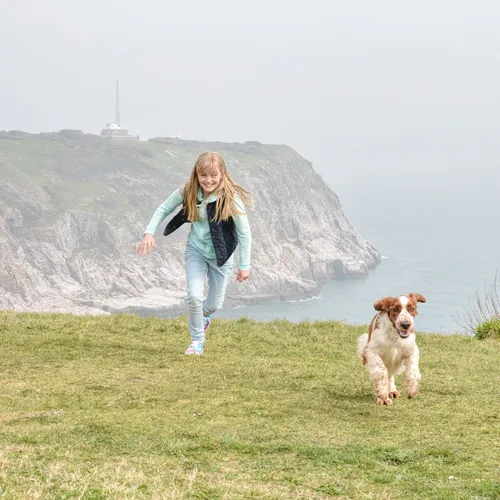 Young girl running with a dog on the clifftop at Berry Head on the South West Coast Path, with the sea and rocky cliffs behind.