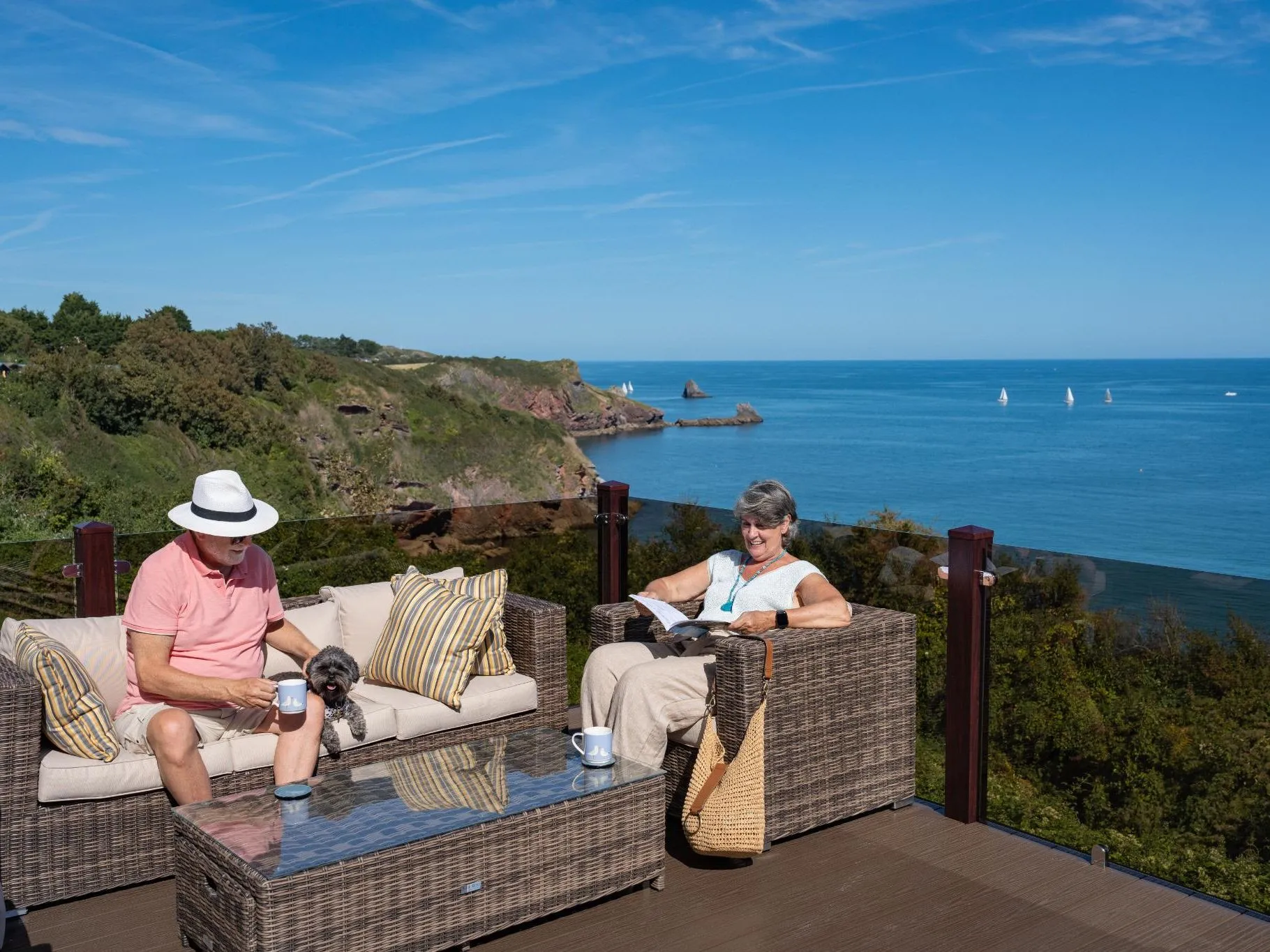 Mature couple relaxing on outdoor decking with a small dog, overlooking the Devon coastline and sea