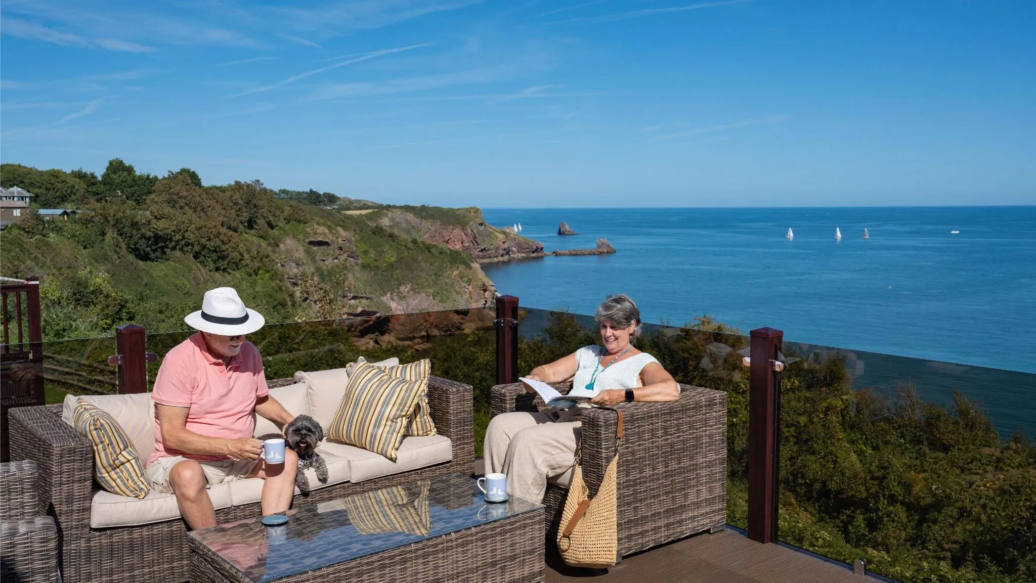 Mature couple relaxing on outdoor decking with a small dog, overlooking the Devon coastline and sea