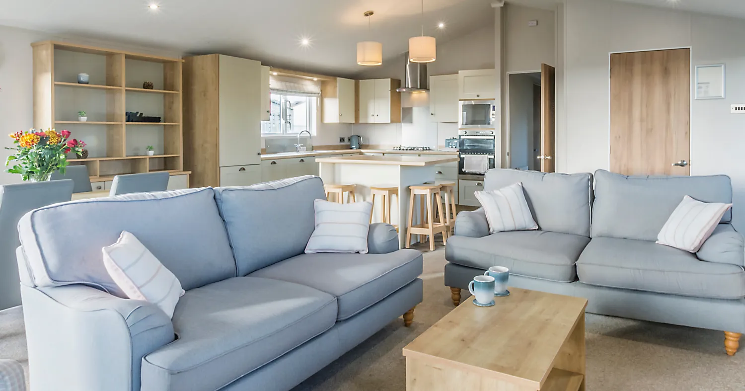 Bright open-plan lodge lounge and kitchen with pale blue sofas, wooden coffee table and breakfast bar stools