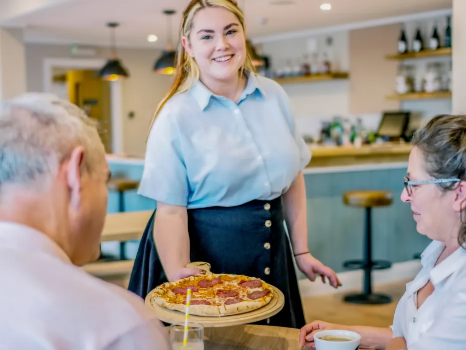 A staff member serving a pizza to residents in a bright, welcoming café area.