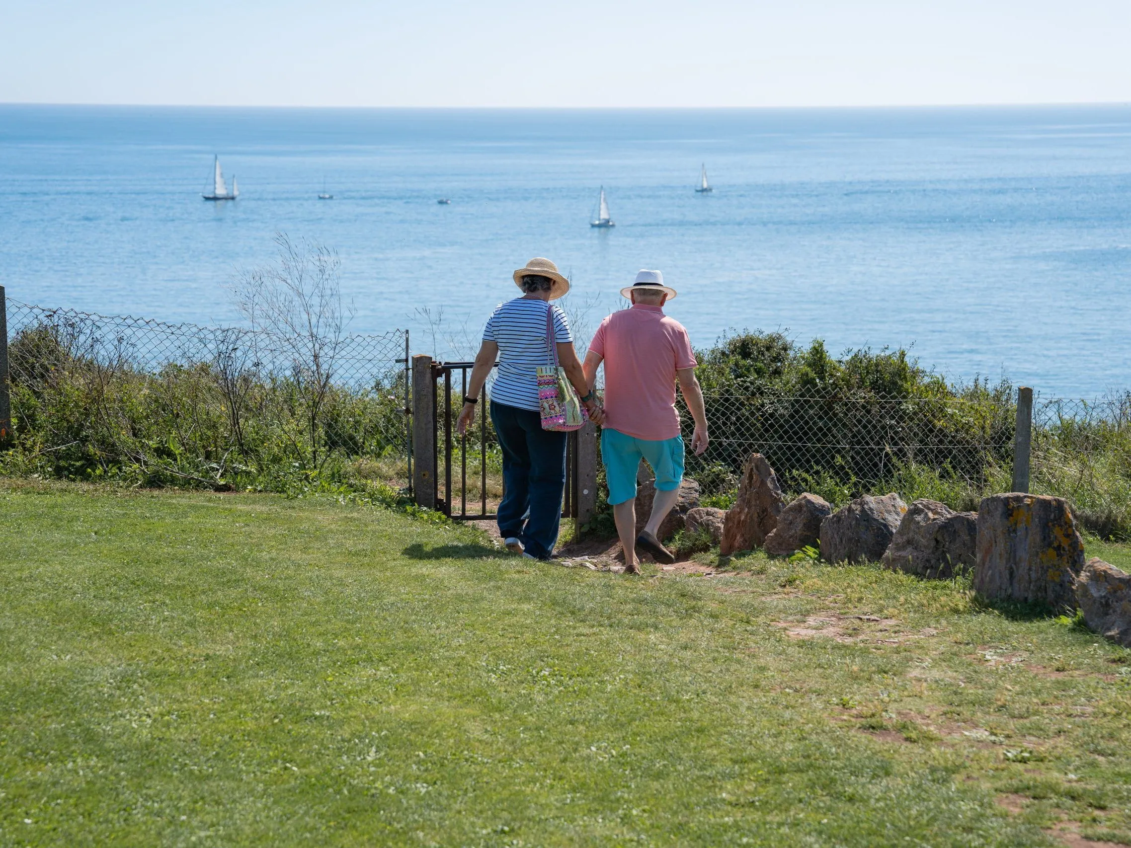 Couple walking through a garden gate towards a coastal view with sailing boats on a calm blue sea