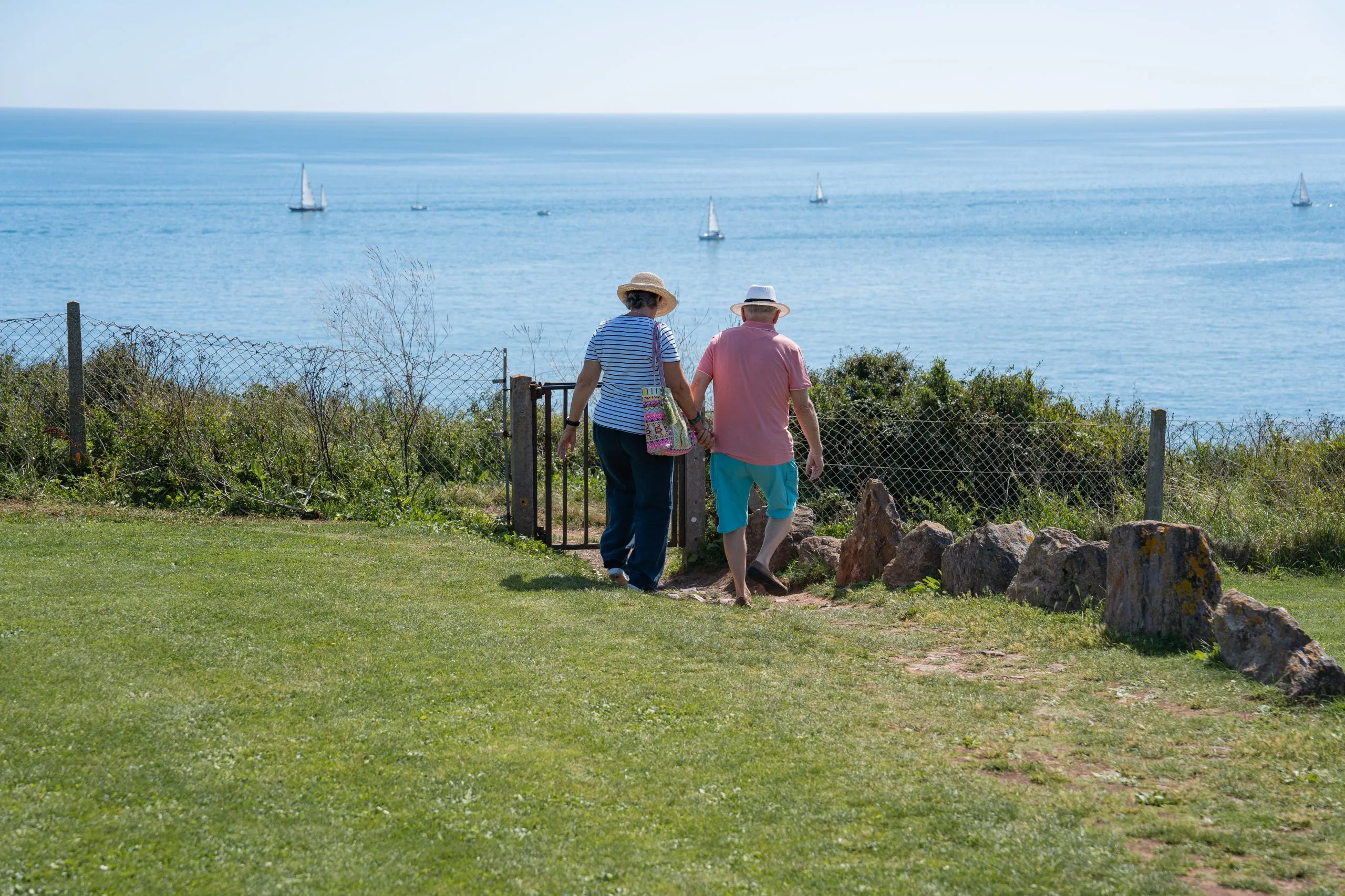 Couple walking through a garden gate towards a coastal view with sailing boats on a calm blue sea