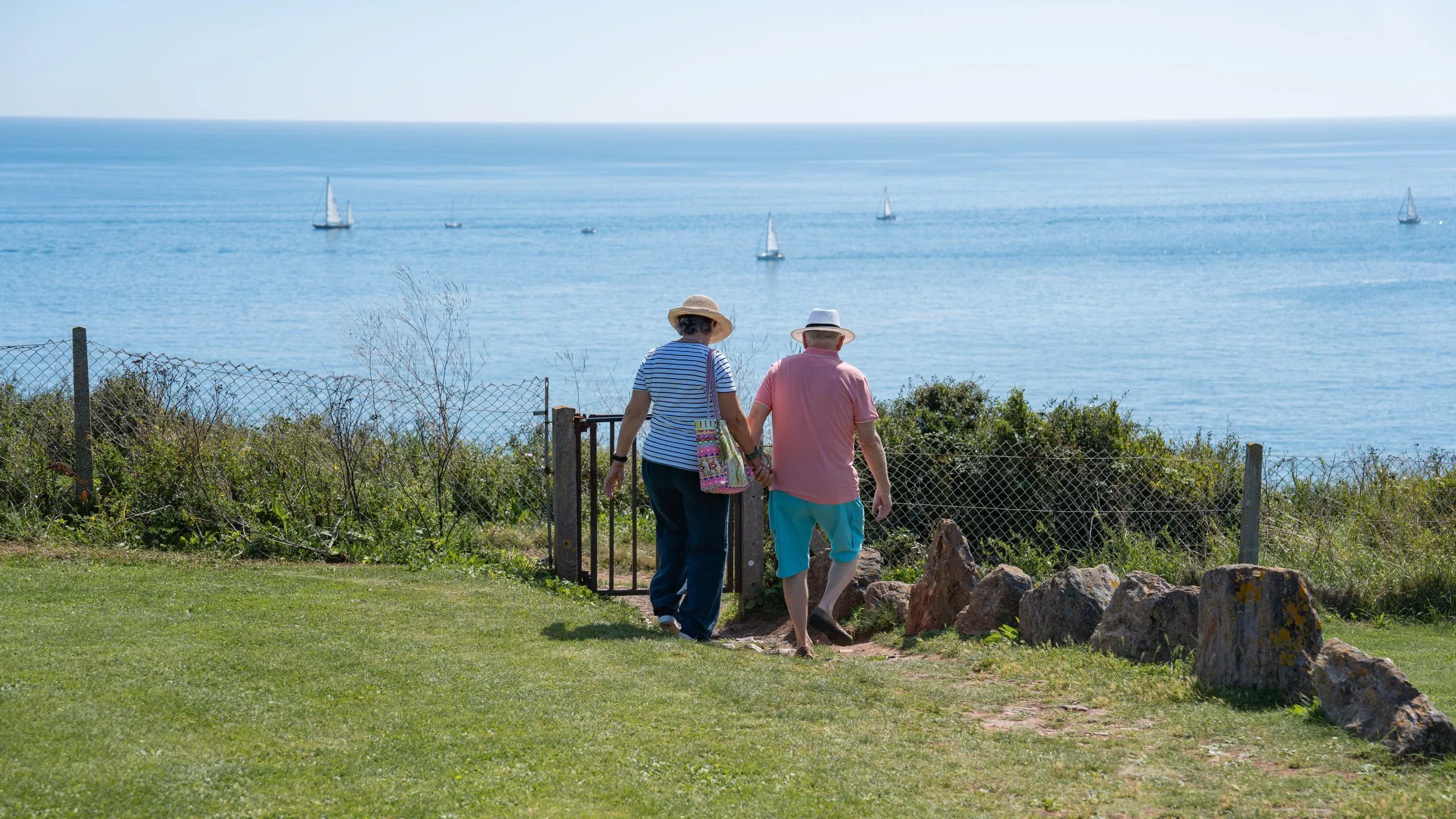 Couple walking through a garden gate towards a coastal view with sailing boats on a calm blue sea