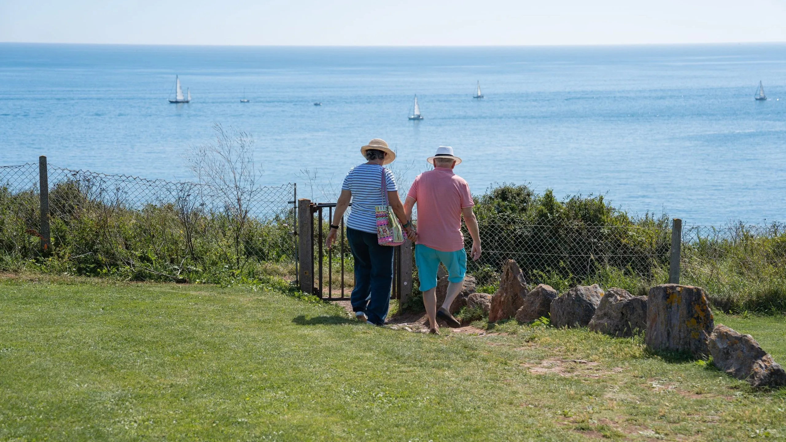 Couple walking through a garden gate towards a coastal view with sailing boats on a calm blue sea