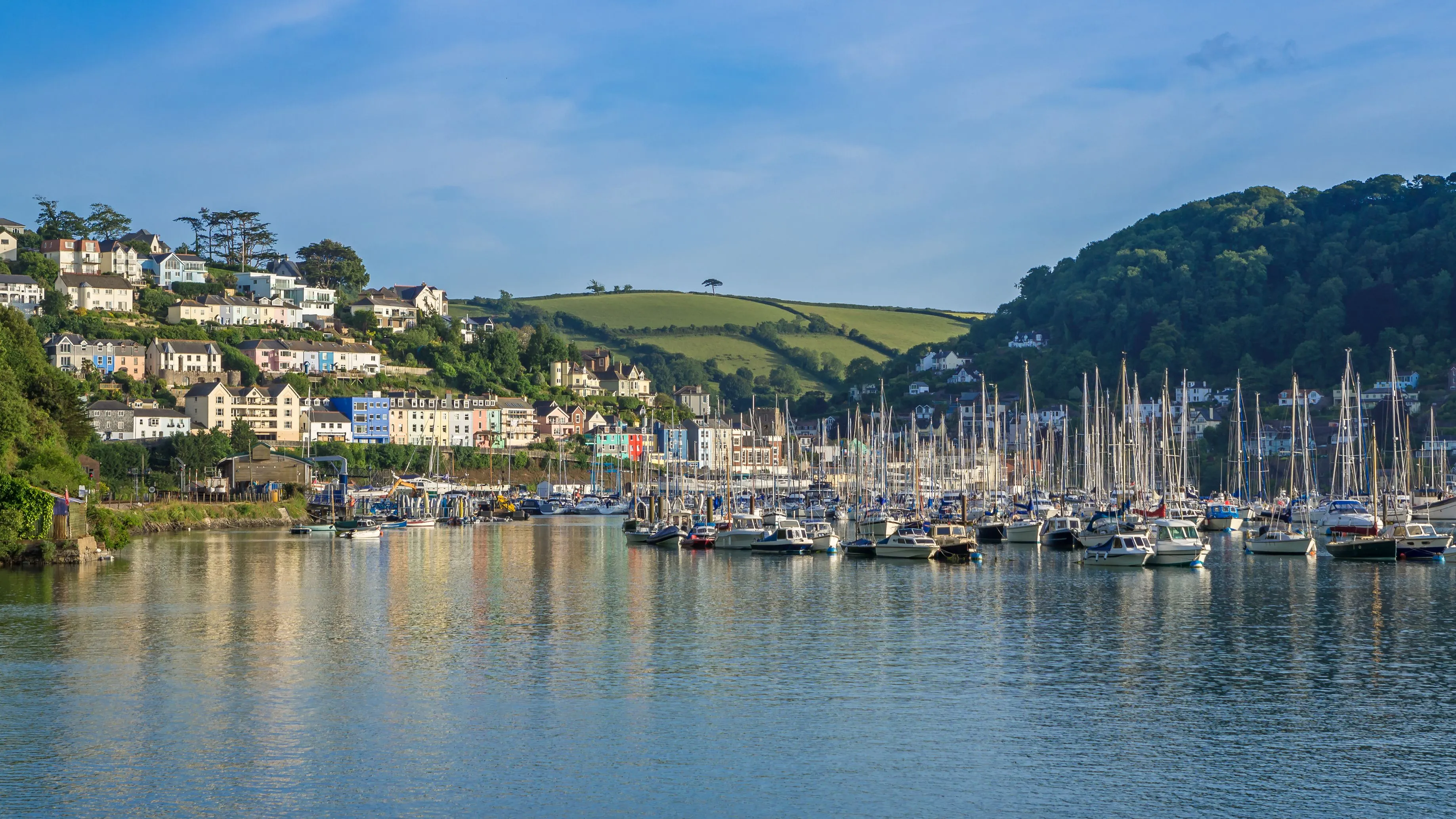 Harbour filled with sailing boats beneath colourful hillside houses and green countryside