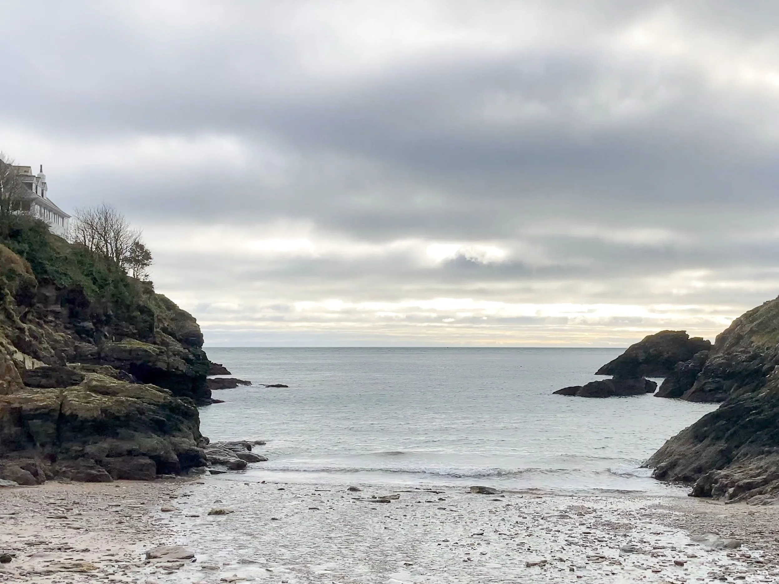 Small rocky cove with a pebbly beach, calm sea and cloudy sky, with a house perched on the left cliff.