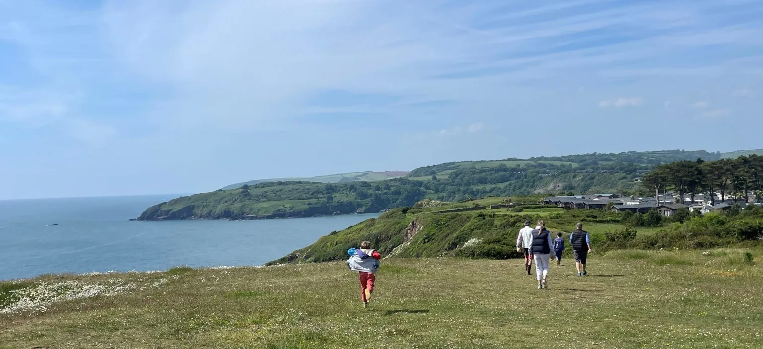 People walking across a grassy coastal headland overlooking the sea and rolling green hills under a blue sky.