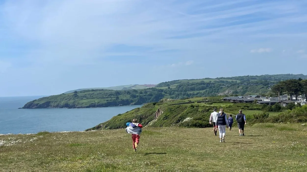 People walking across a grassy coastal headland overlooking the sea and rolling green hills under a blue sky.