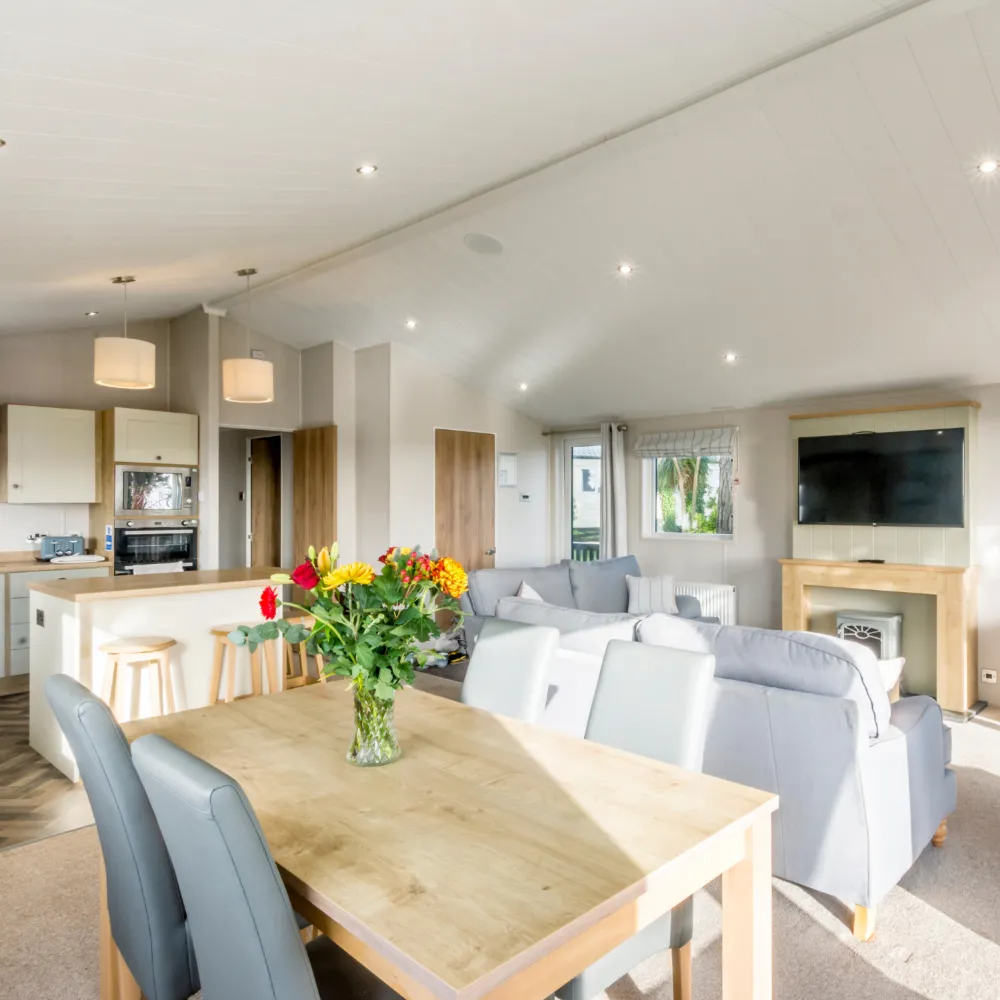 Open-plan lodge dining area with wooden table, grey chairs and sofa, and fitted kitchen