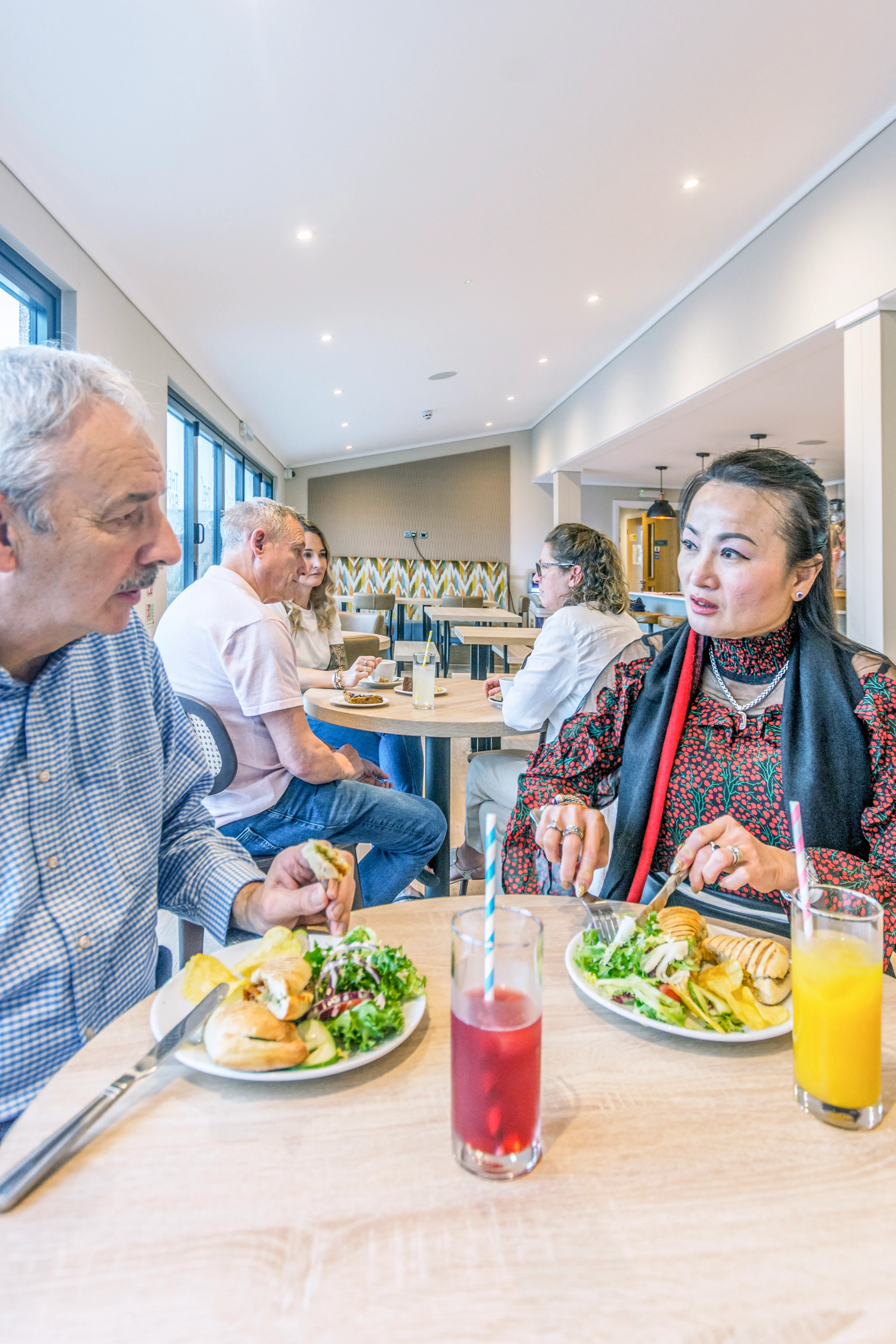 Two people enjoying lunch and conversation at a bright café table with salads, sandwiches and drinks.