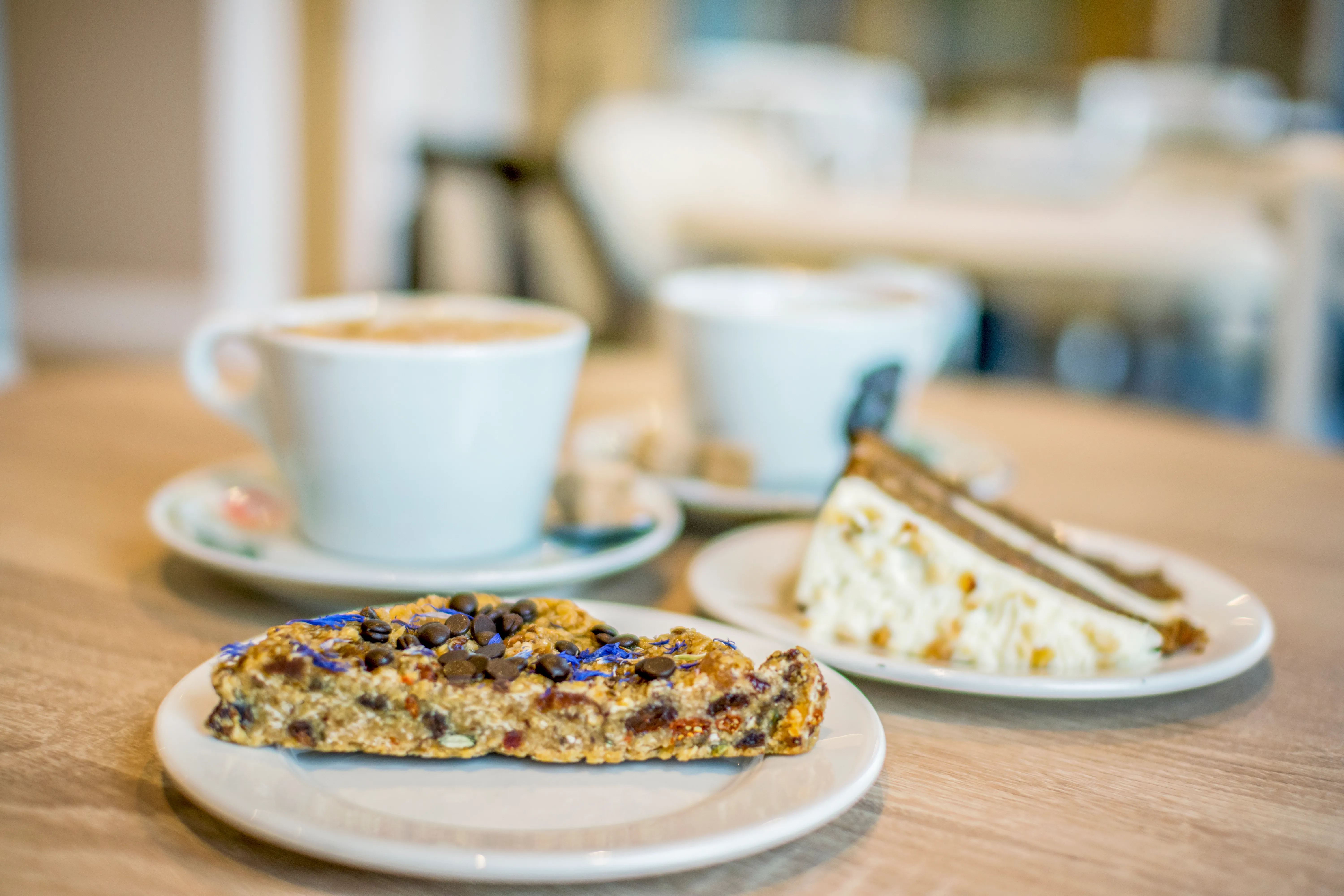 Two cups of coffee with a slice of cake and a fruit flapjack on a café table.