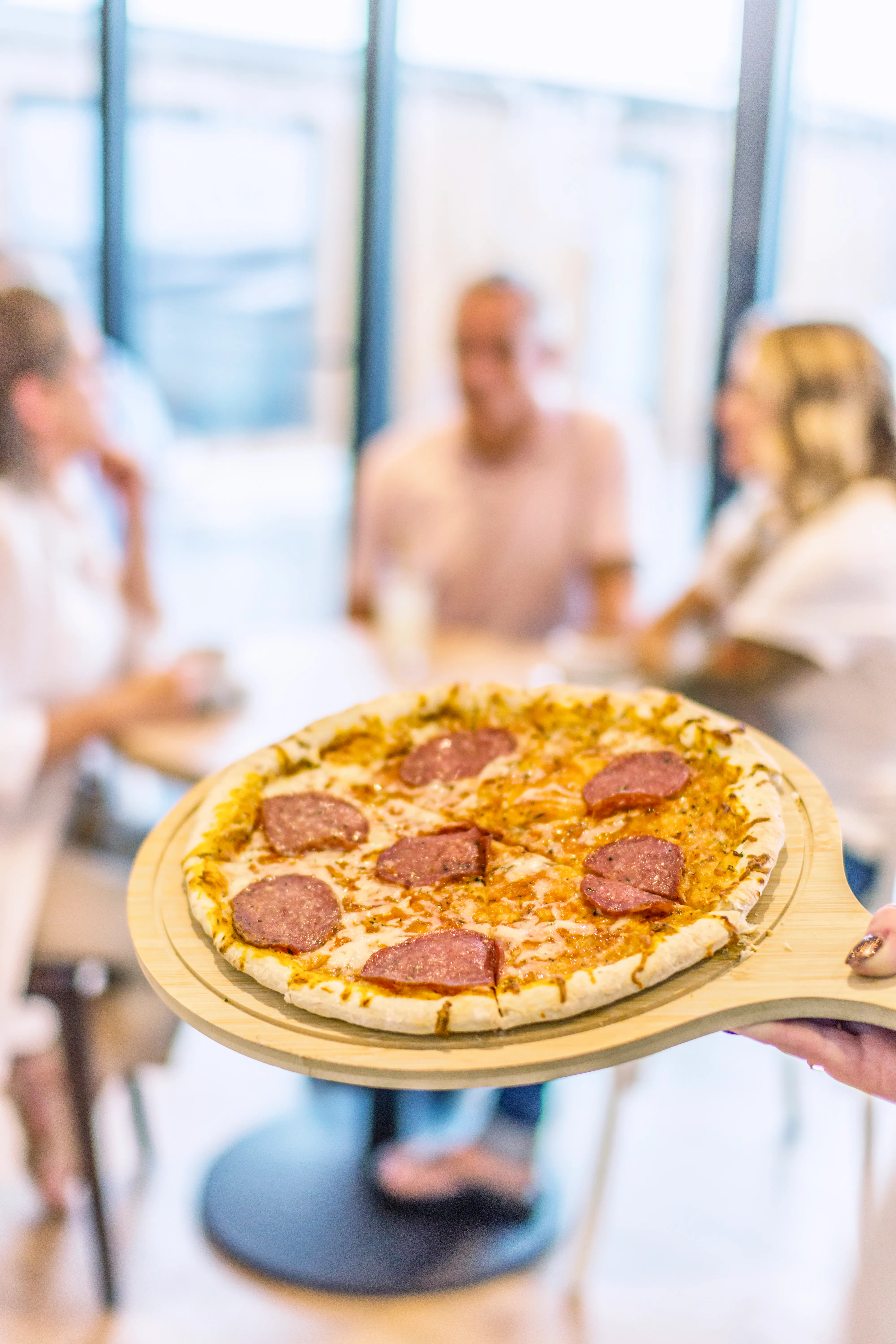 Pepperoni pizza on a wooden serving board with people chatting in the background