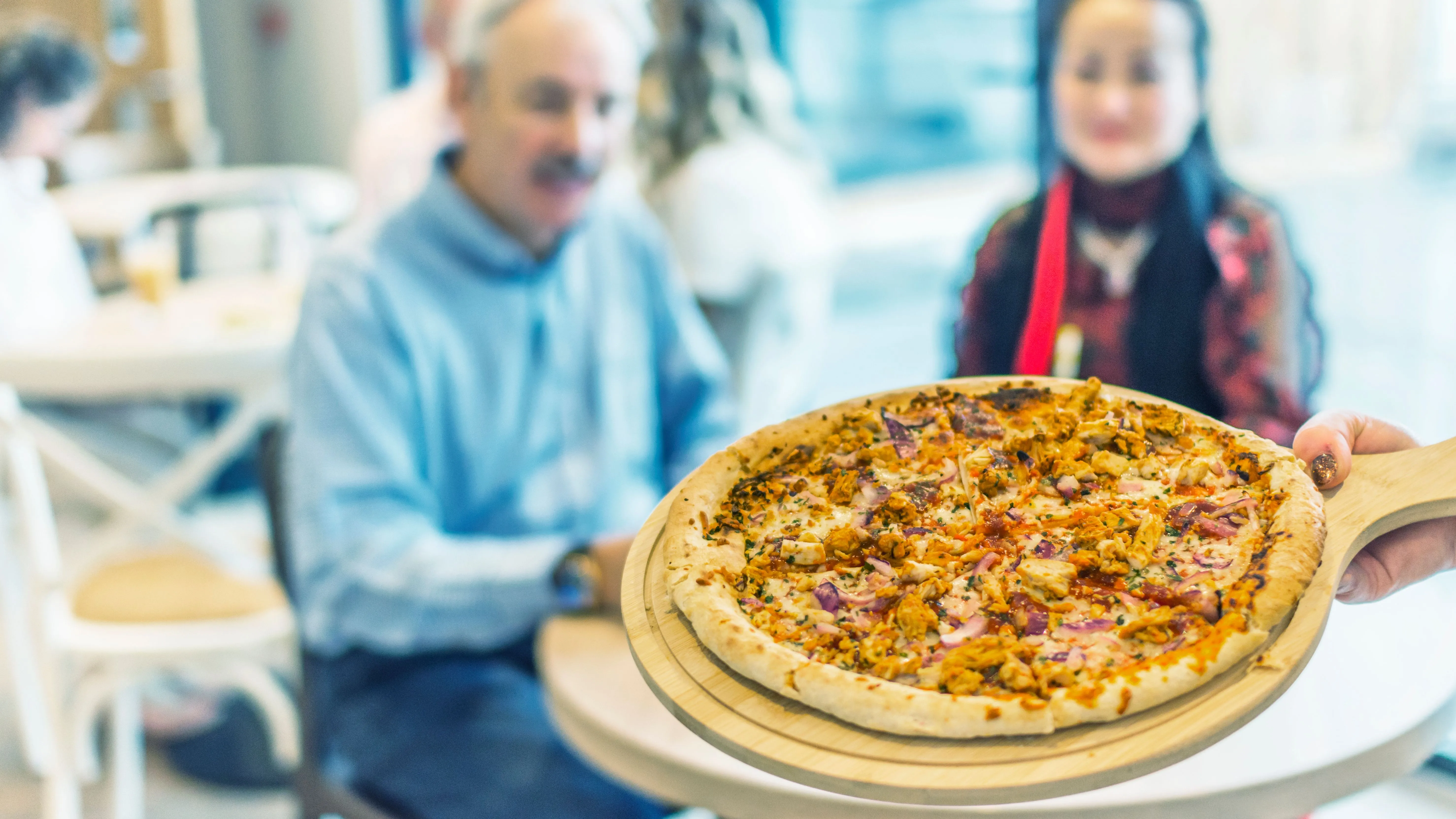 Freshly baked pizza served on a wooden peel in a bright café, with diners seated in the background.