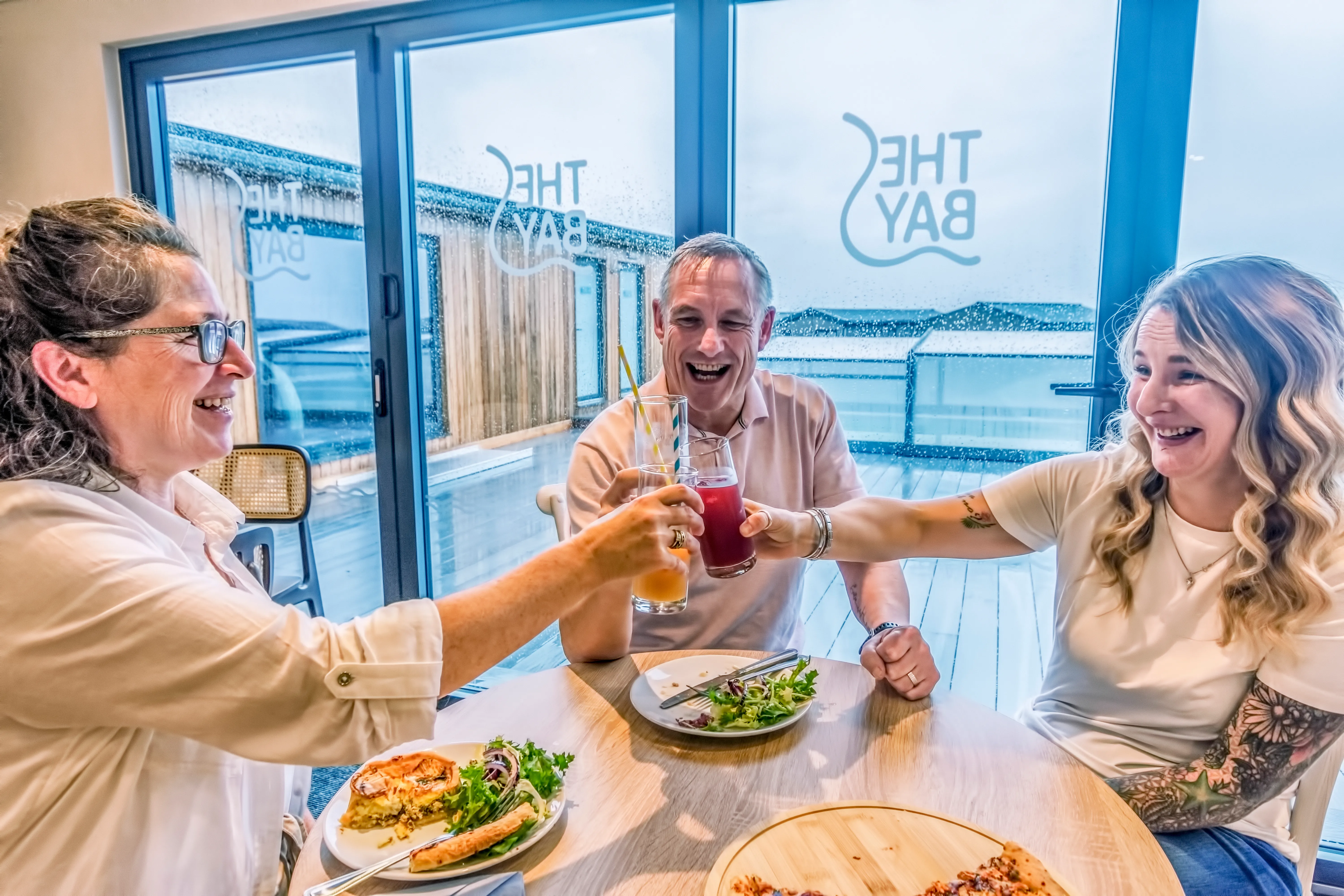 Three people smiling and clinking glasses at a restaurant table beside large windows on a rainy day.
