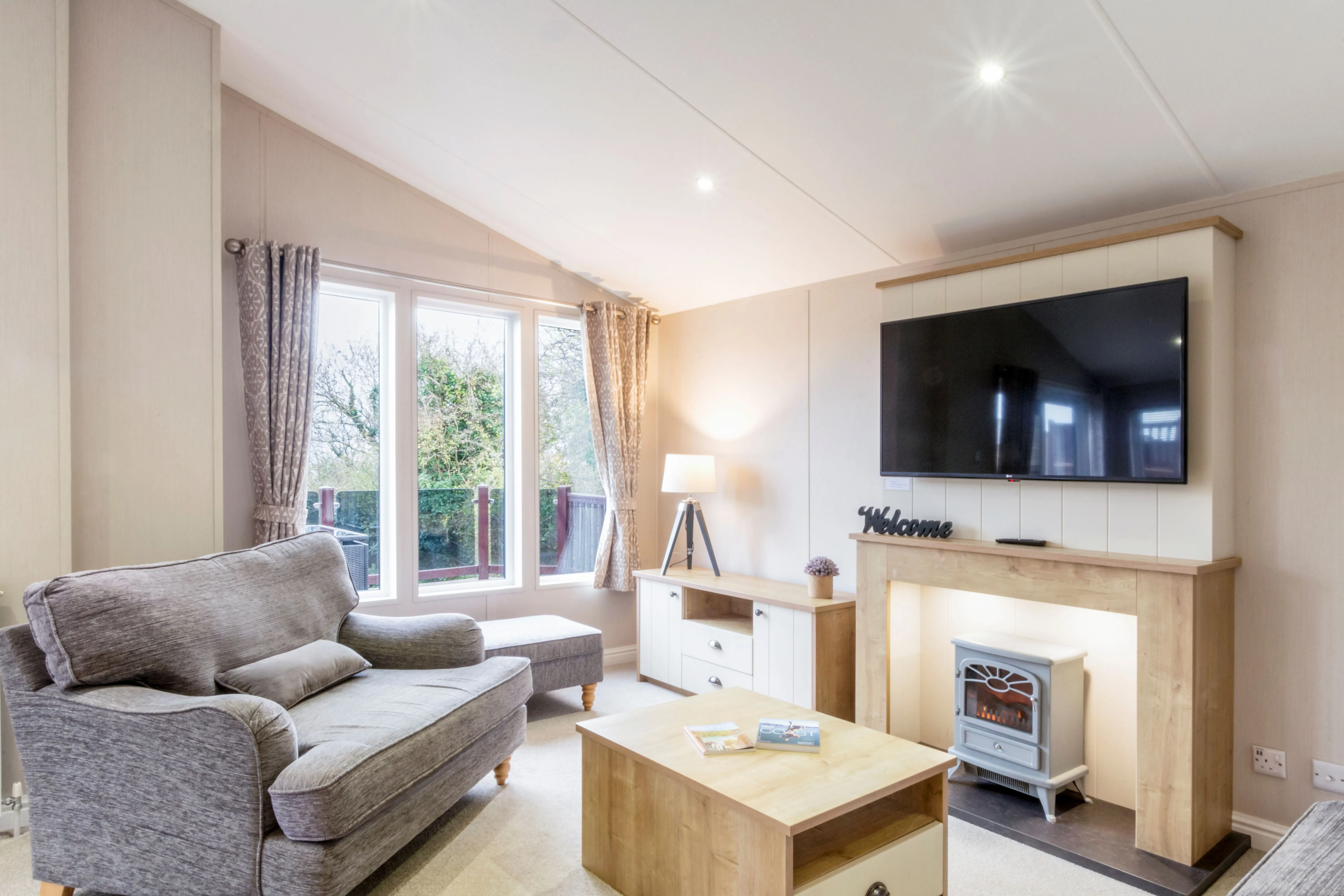 Bright, modern living room with a grey sofa, large windows, wall-mounted television and electric fire.