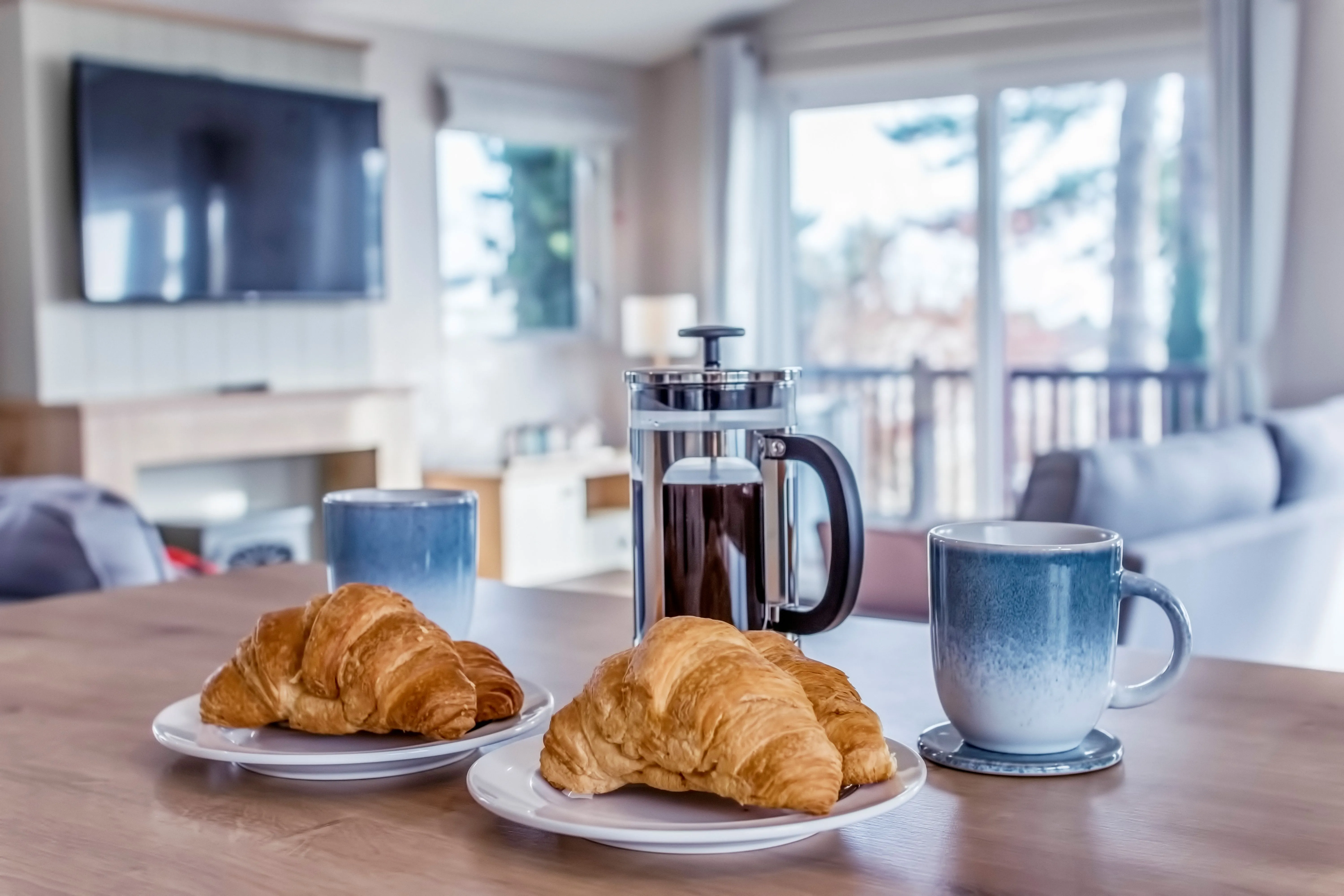 Breakfast table with croissants, coffee press and mugs in a bright cosy living room