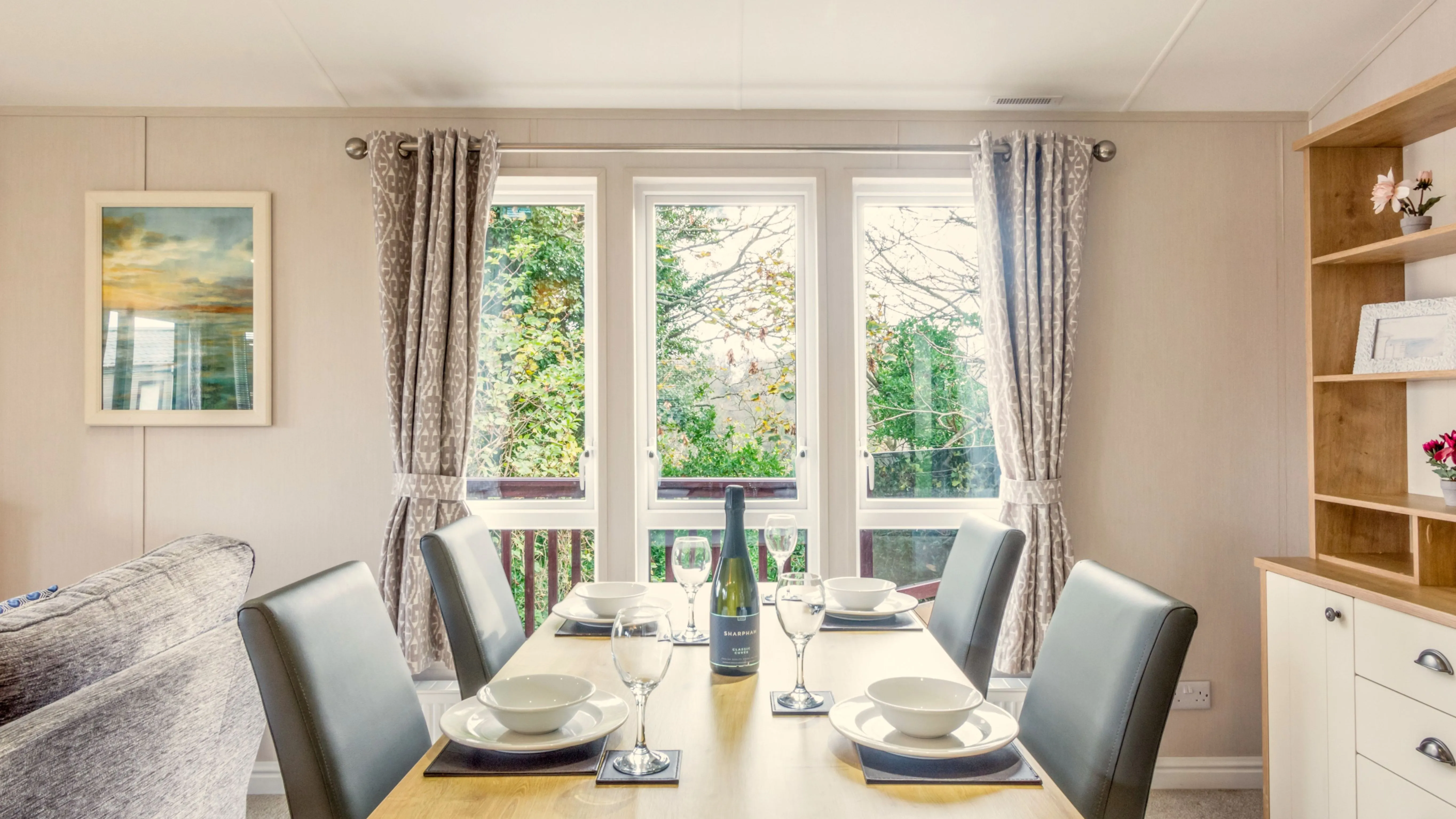 Dining area with a wooden table set for four, upholstered chairs, and large windows with patterned curtains.