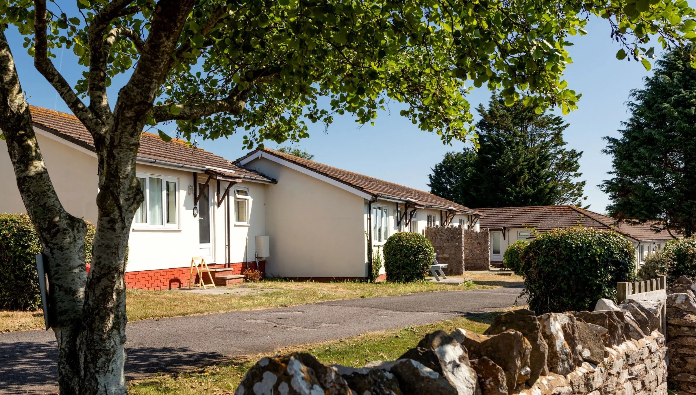 Single-storey holiday bungalows along a quiet lane with a stone wall and leafy tree in the foreground