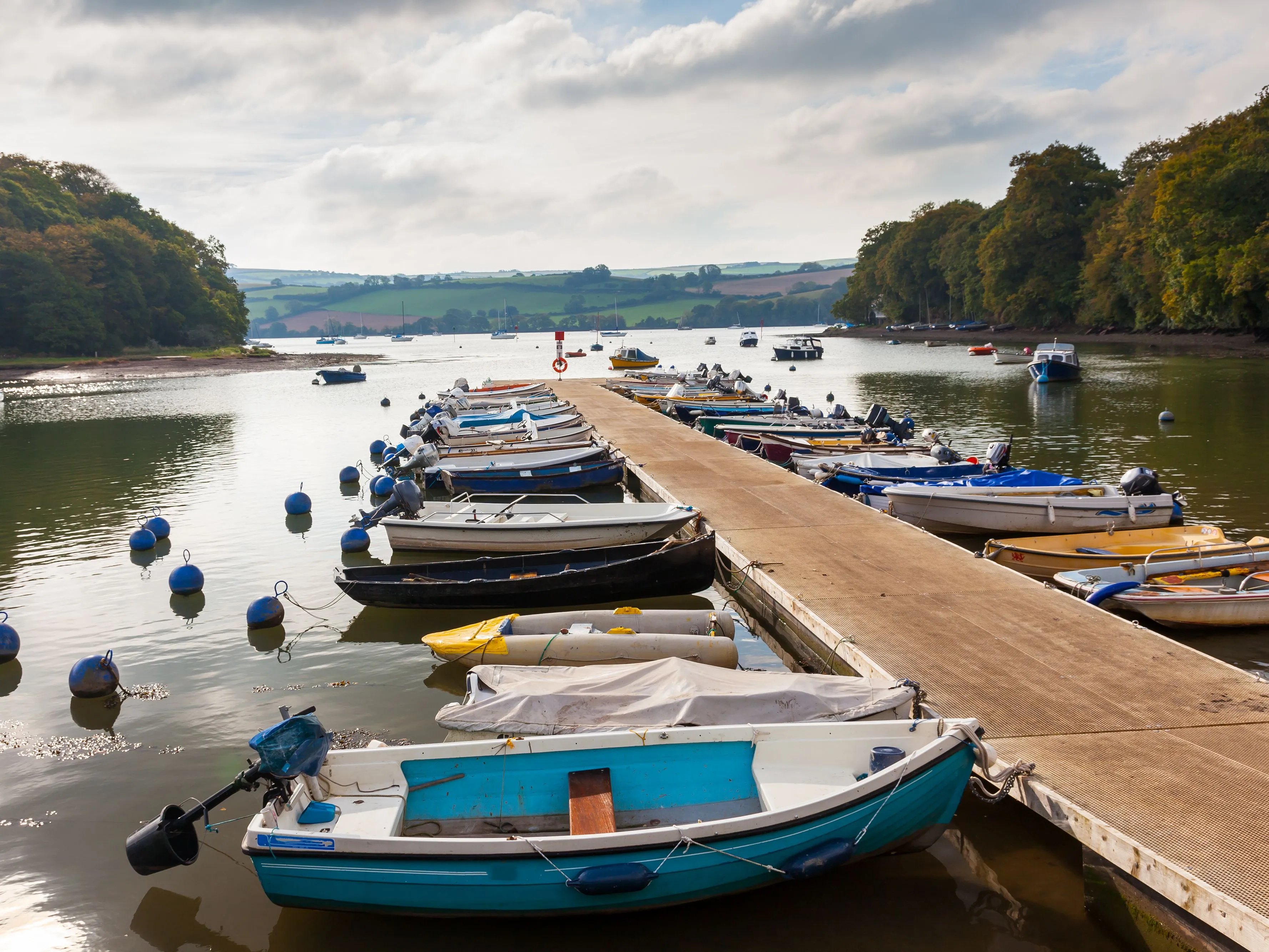 Small boats moored alongside a floating pontoon on a calm river estuary surrounded by wooded hills and countryside.