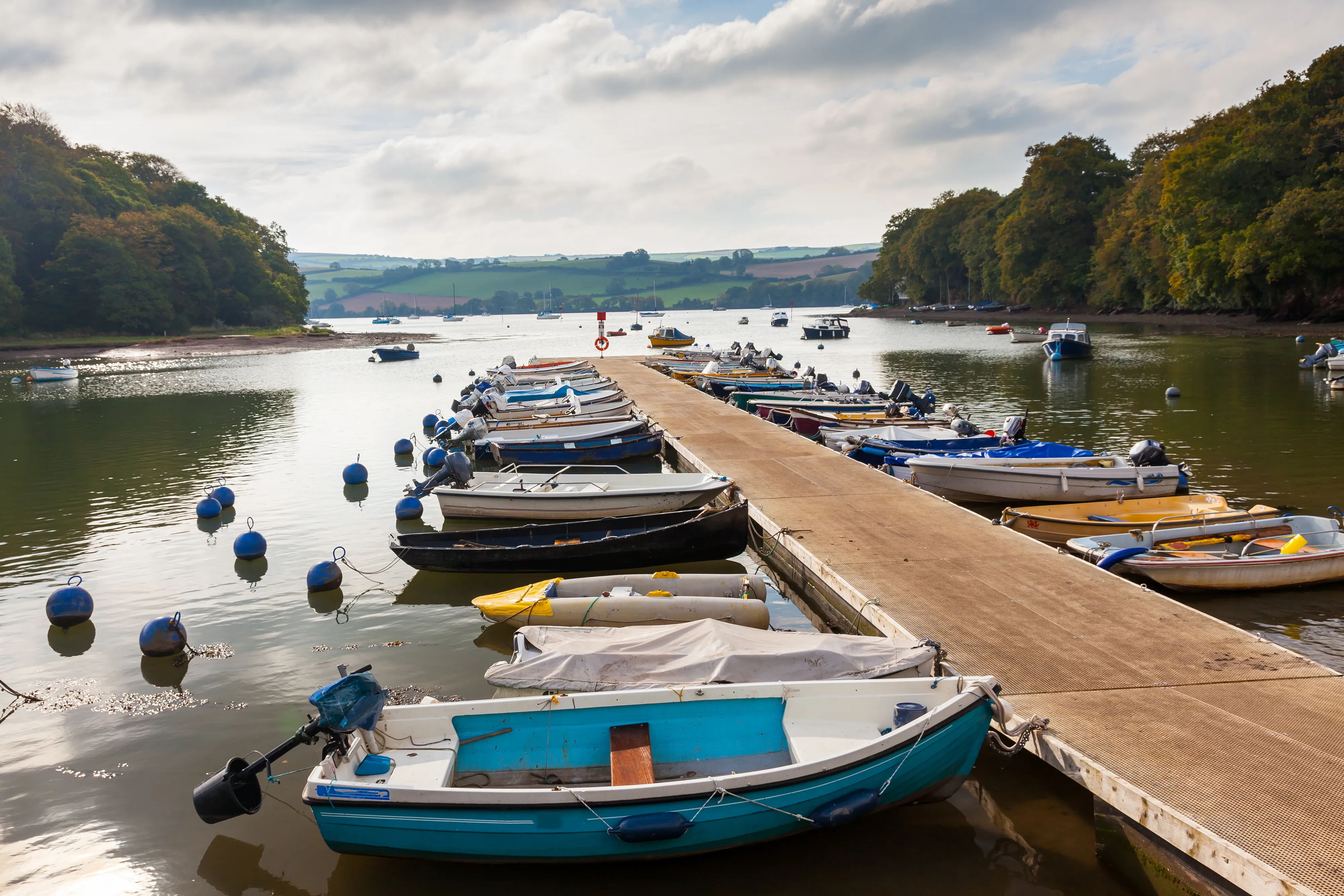 Small boats moored alongside a floating pontoon on a calm river estuary surrounded by wooded hills and countryside.