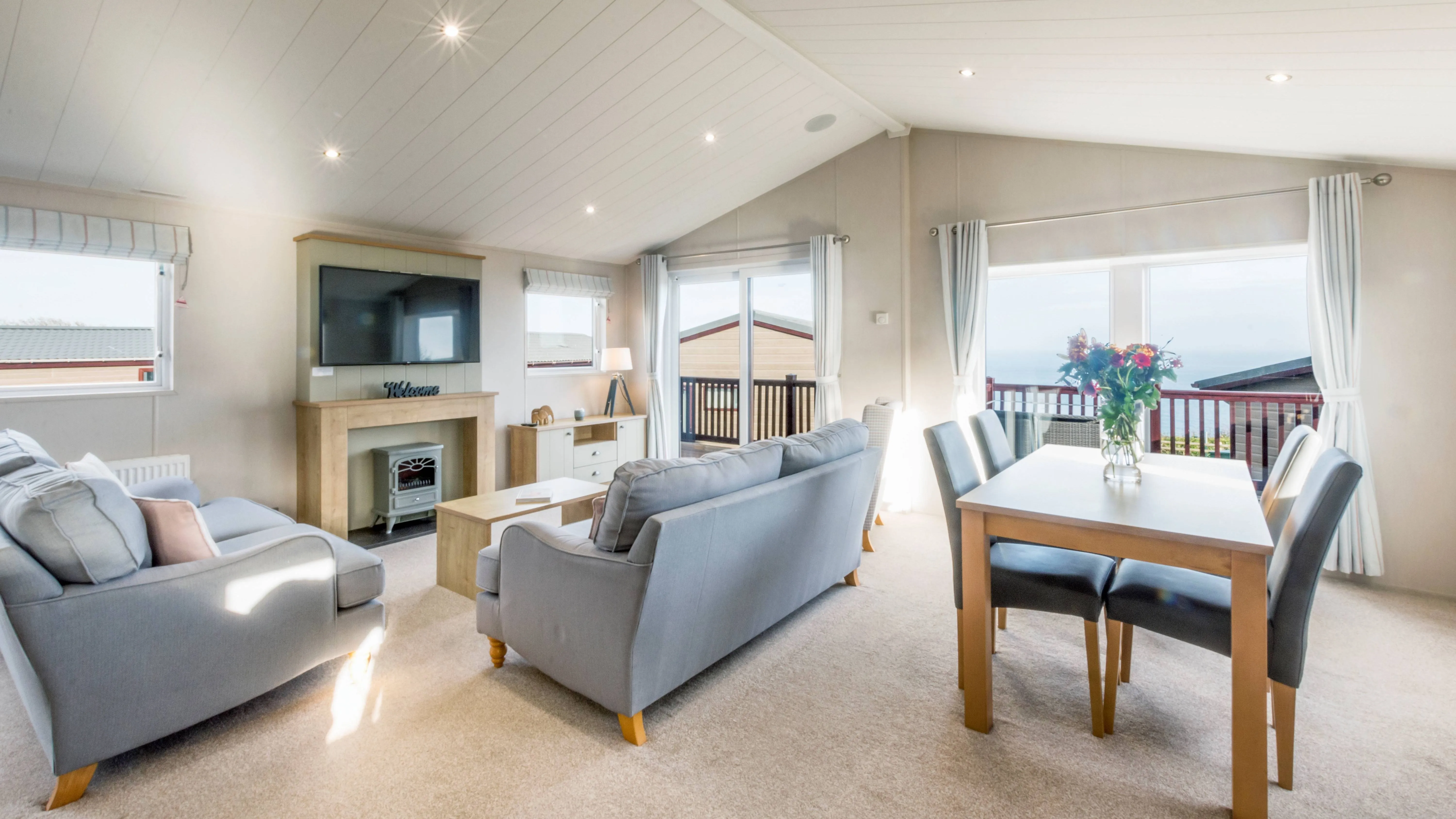 Bright and spacious holiday lodge living area with sofas, dining table, fireplace, TV and balcony views
