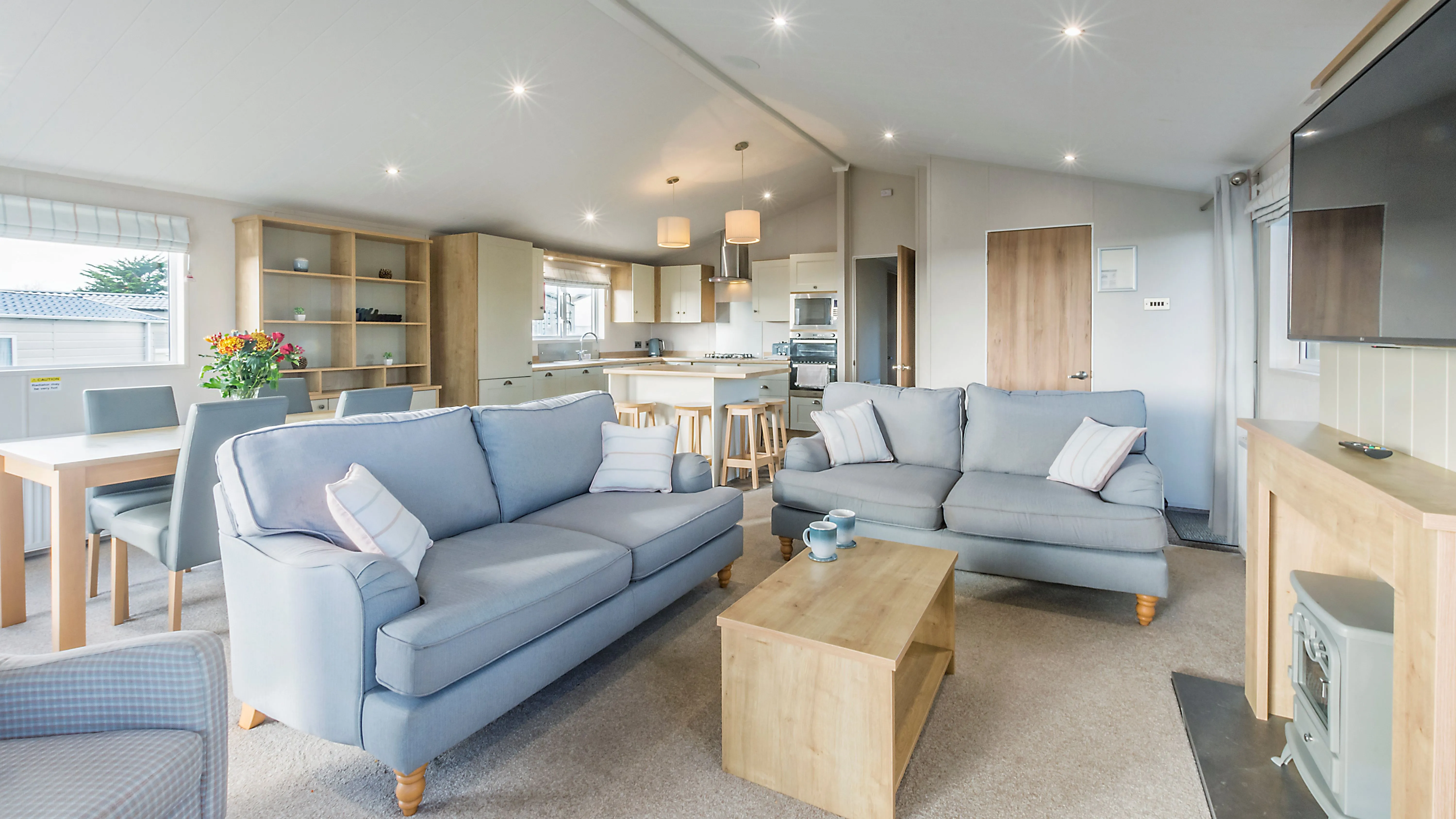 Spacious open-plan living area inside Landscove Lodge with grey sofas, dining table, modern kitchen and wall-mounted television