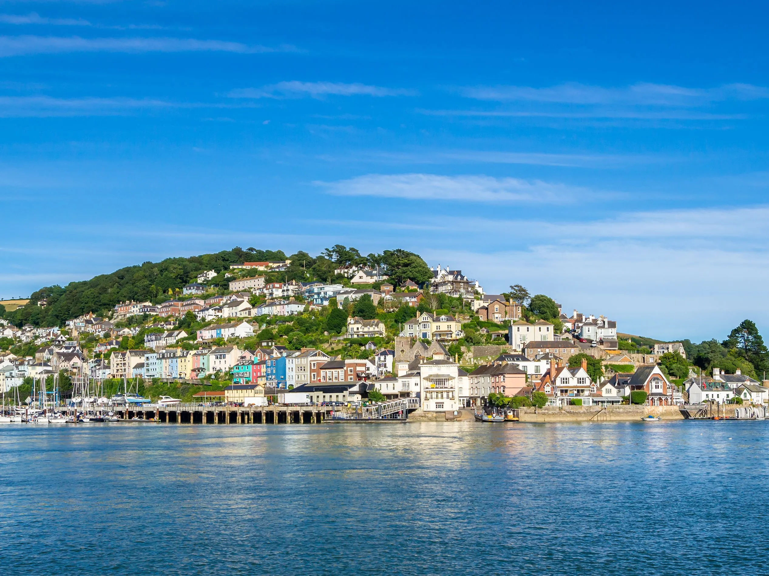 Harbour town with colourful houses climbing a green hillside beside calm blue water under a bright sky