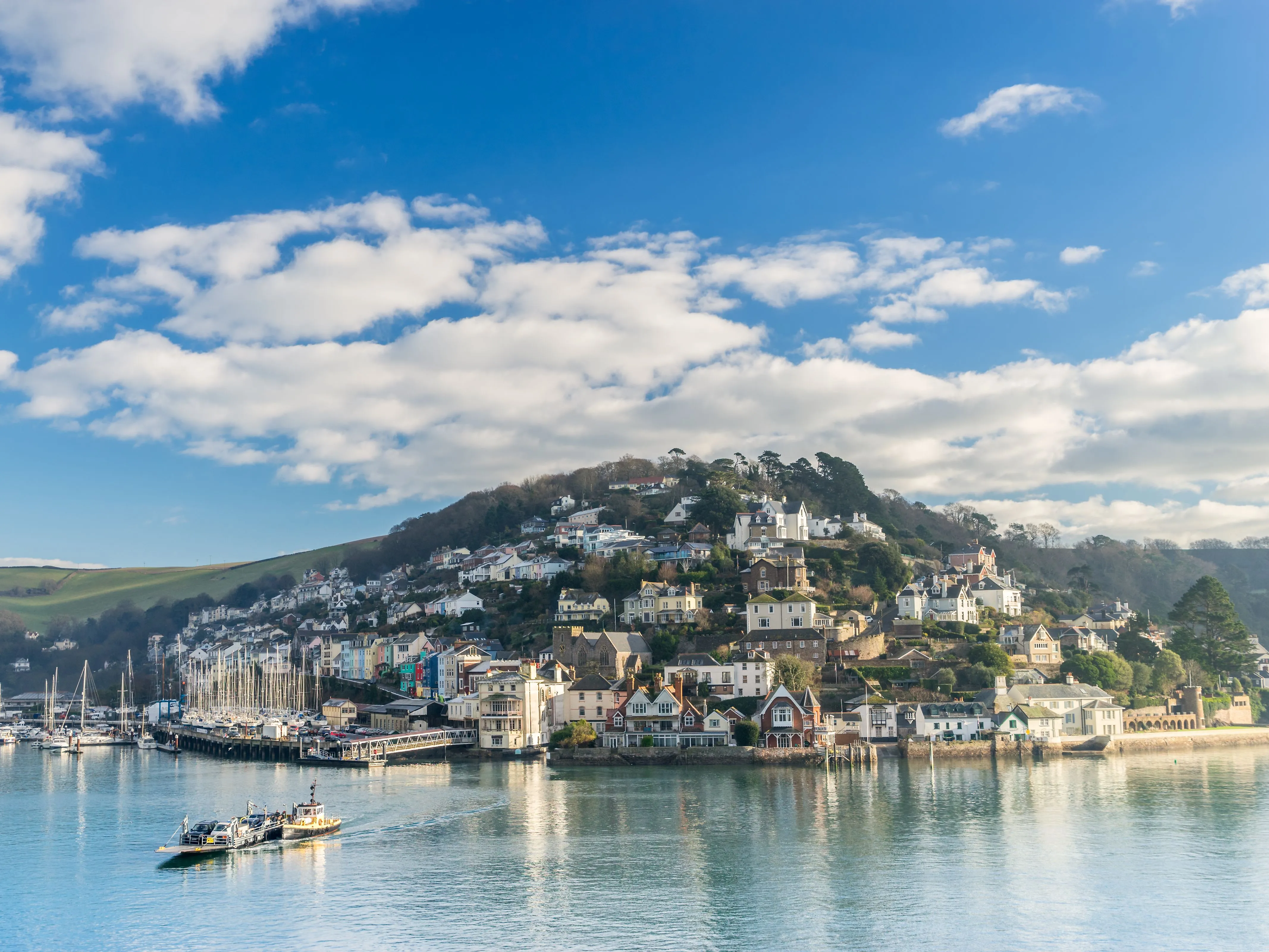 Harbour town with hillside houses, moored yachts and a small ferry on calm water beneath a bright blue sky with scattered clouds