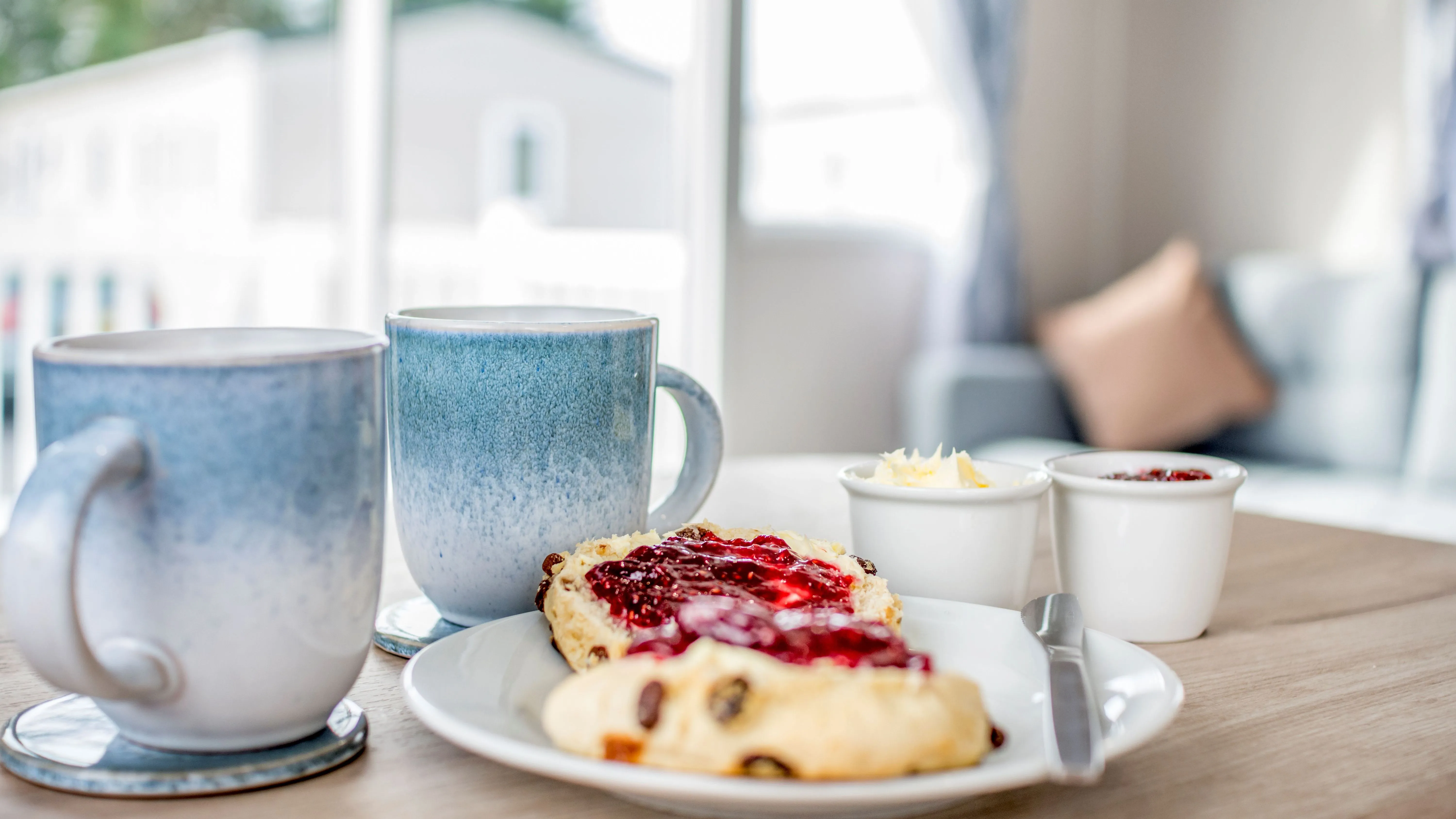 Two mugs beside a fruit scone with jam and cream on a plate in a bright, cosy caravan lounge