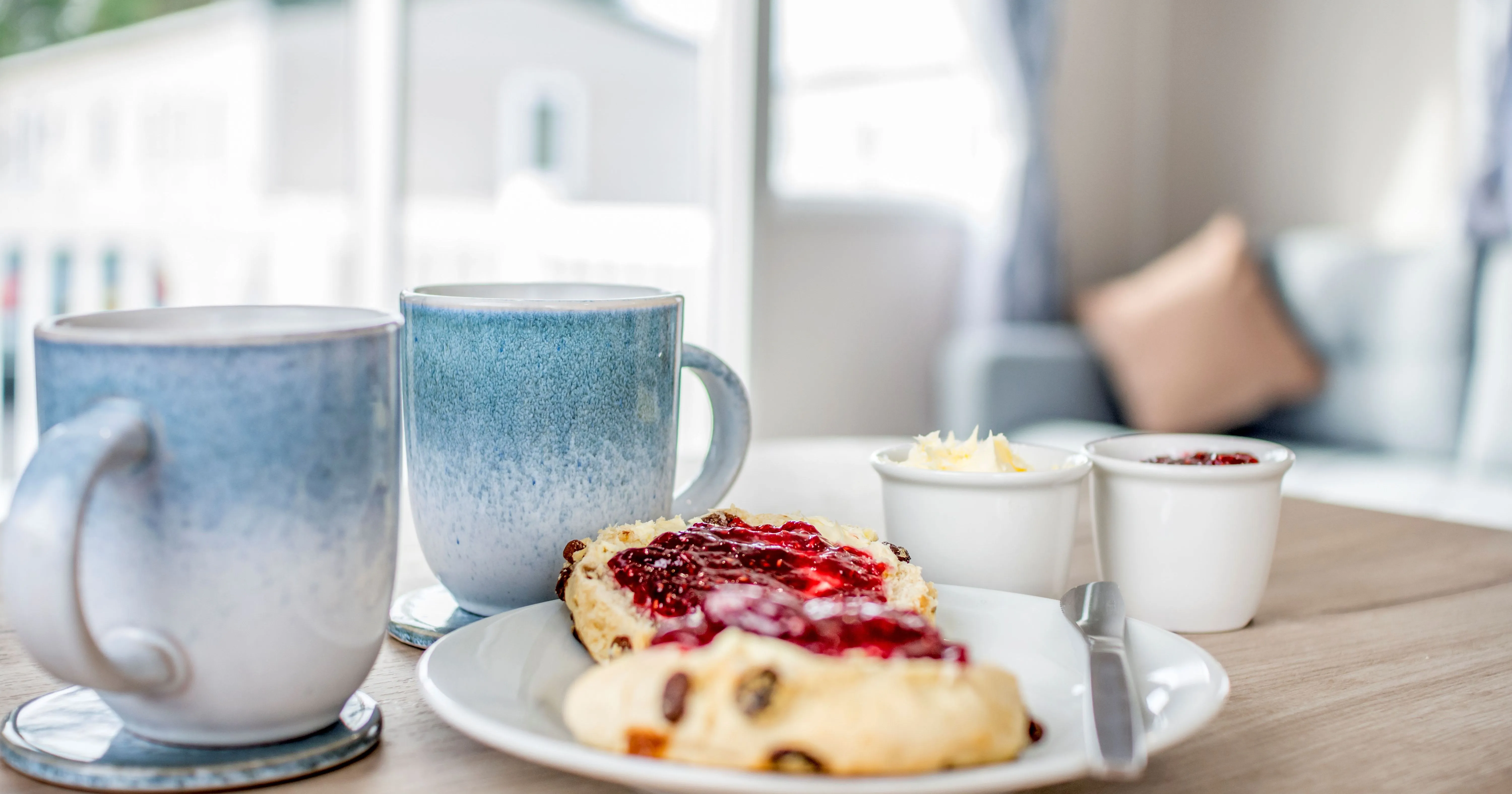 Two mugs beside a fruit scone with jam and cream on a plate in a bright, cosy caravan lounge