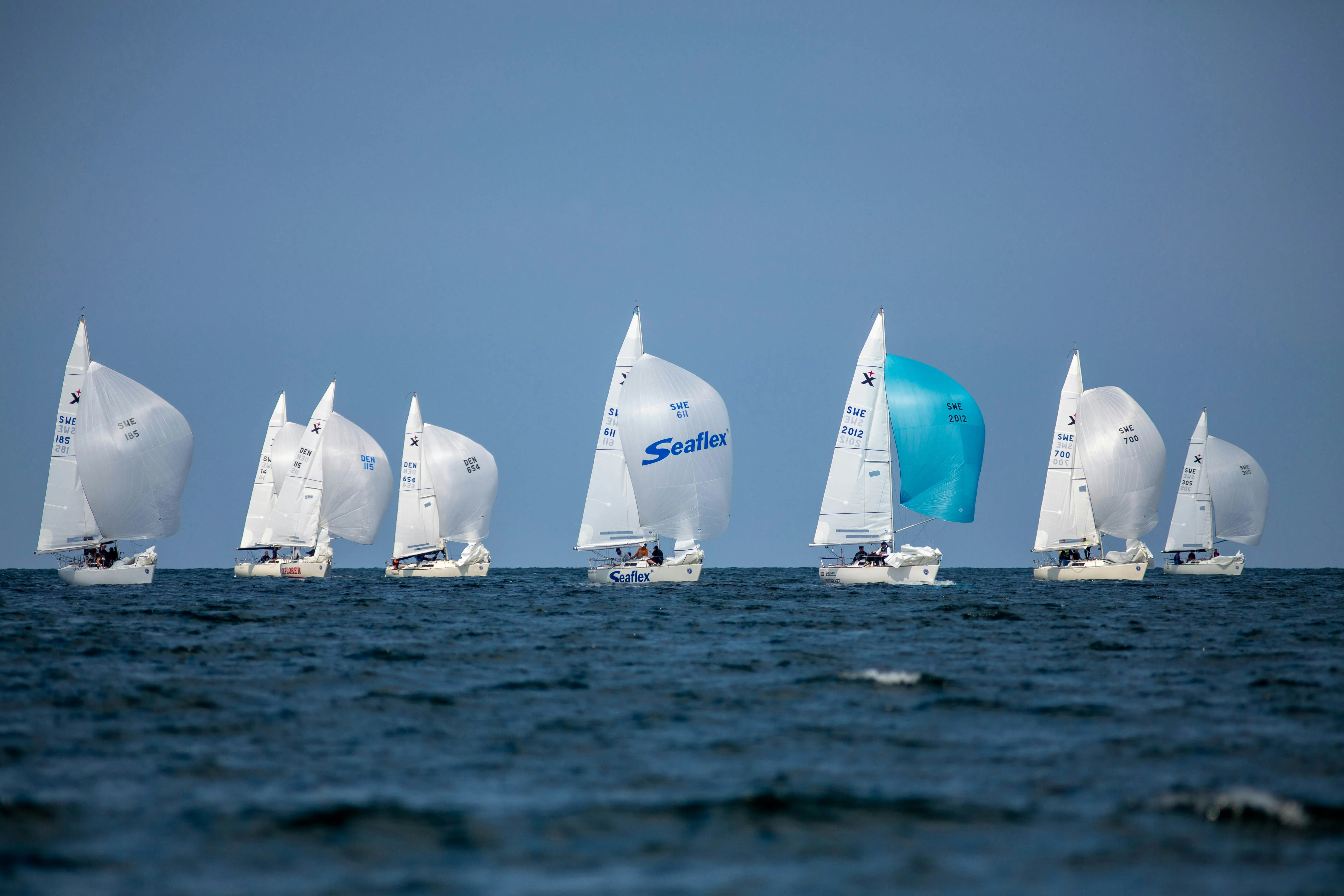 A group of sailing boats racing across the sea with large white and blue spinnakers under a clear sky.